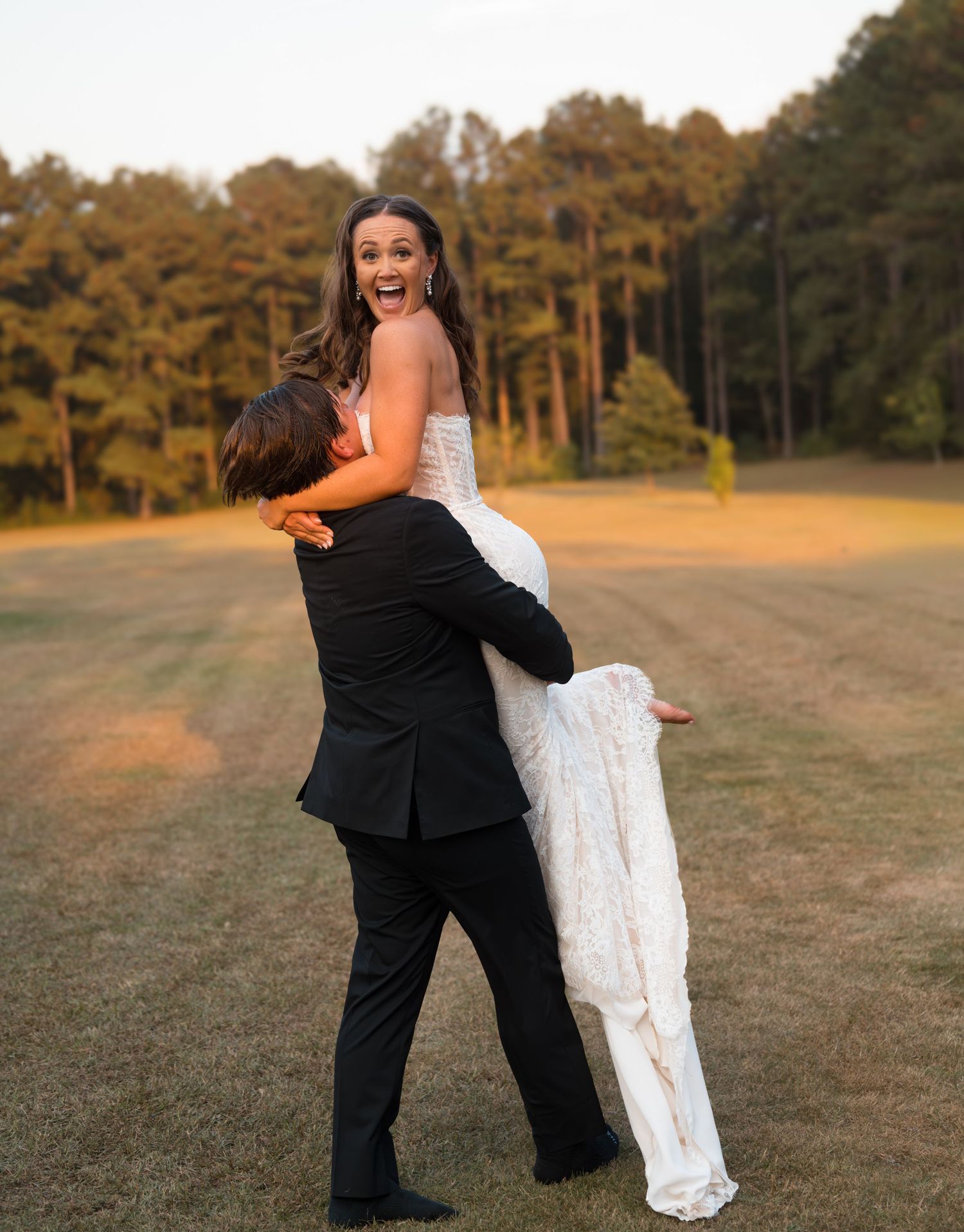 Joyful wedding couple embracing on grass field with pine forest backdrop.
