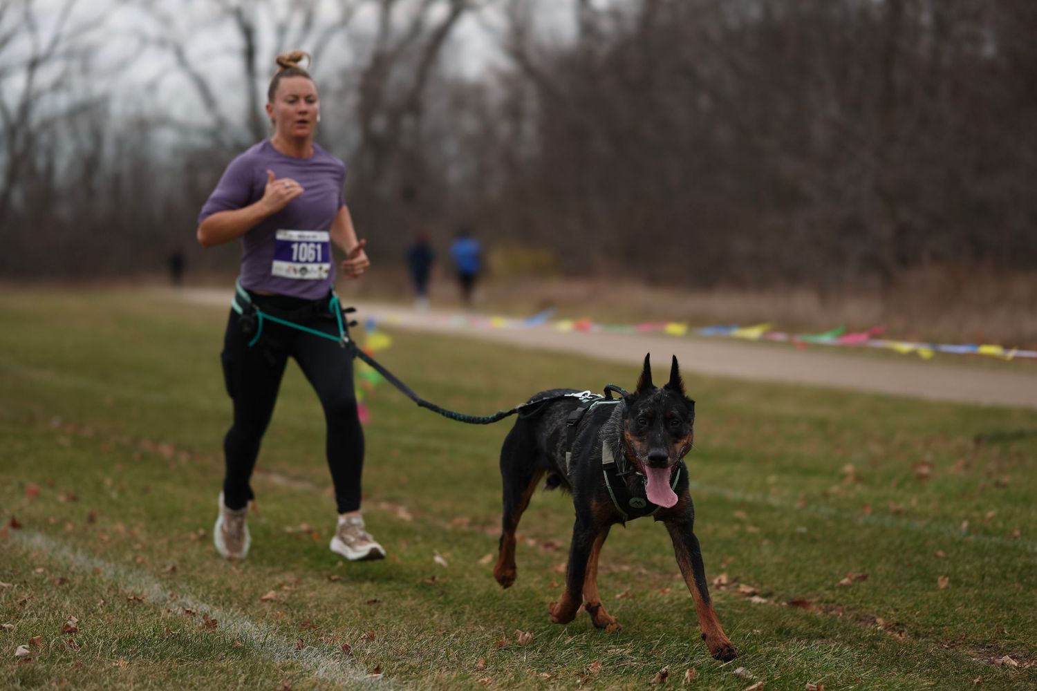 A person and a black Doberman dog running together on a trail in an autumn landscape.