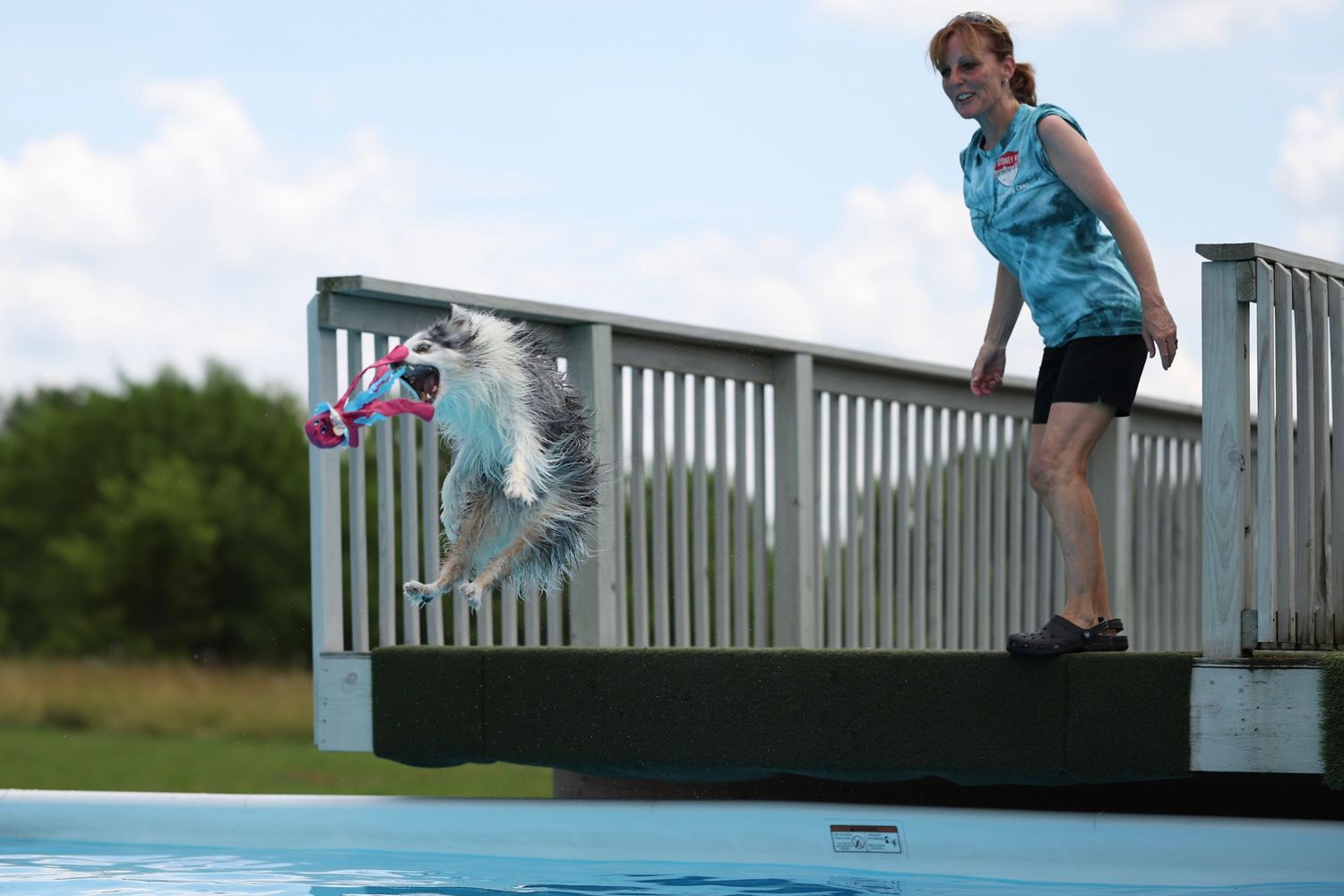 A child in a blue shirt climbs over a white railing on a wooden deck overlooking a lake.