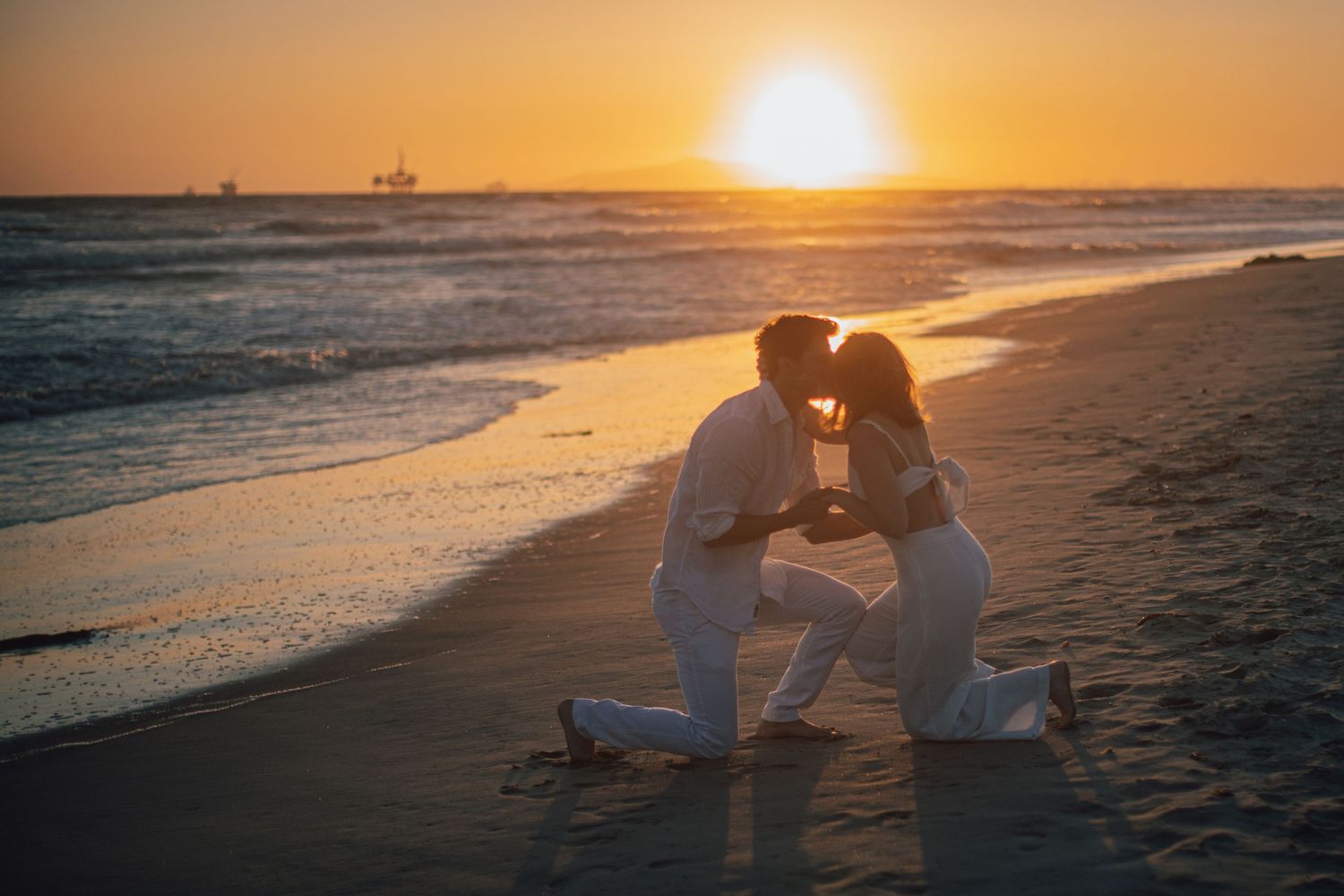 Silhouette of couple kneeling on beach at golden sunset with ships on horizon.