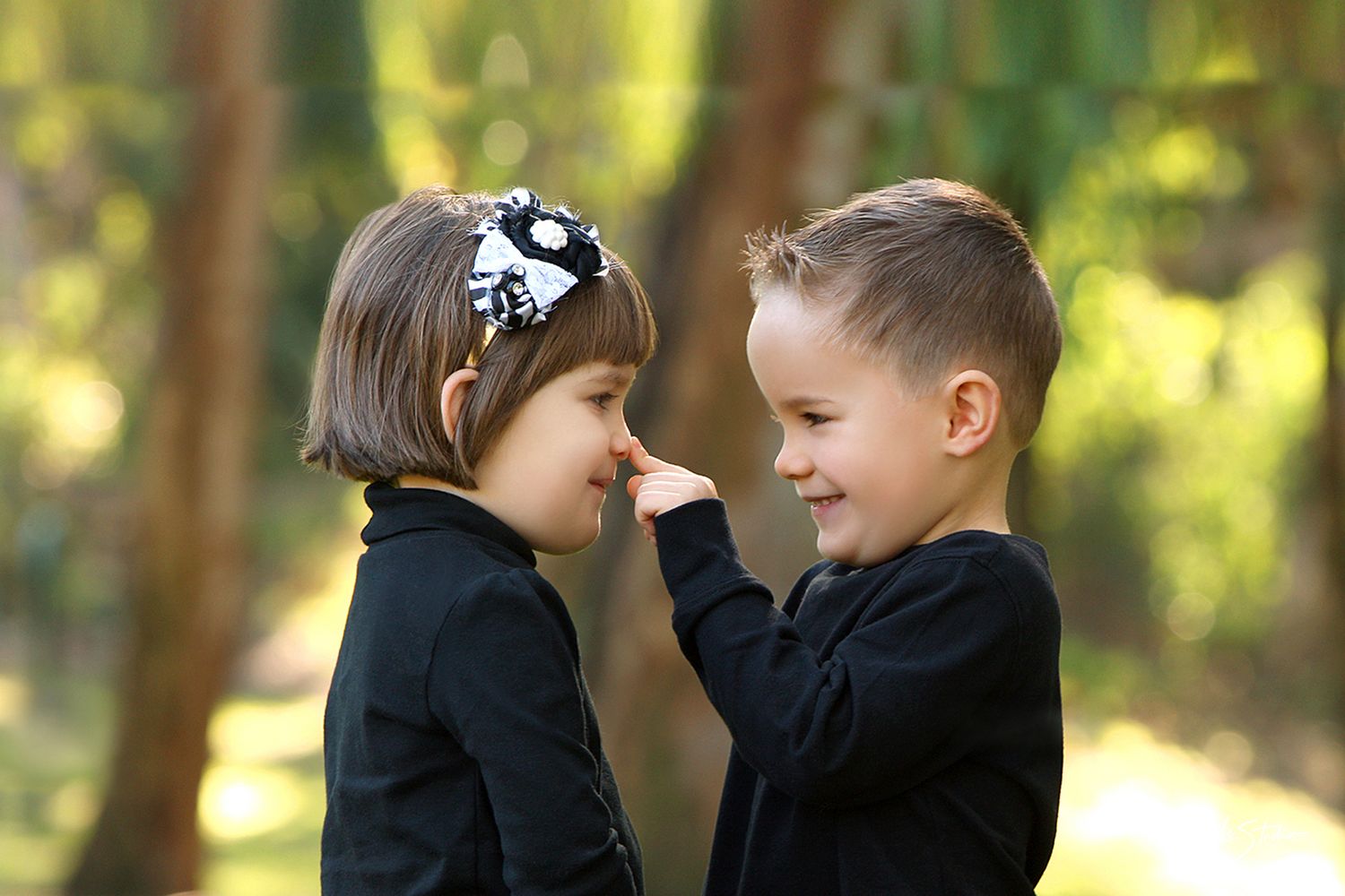 Two children in black sweaters share a playful moment together in a sunlit forest setting.