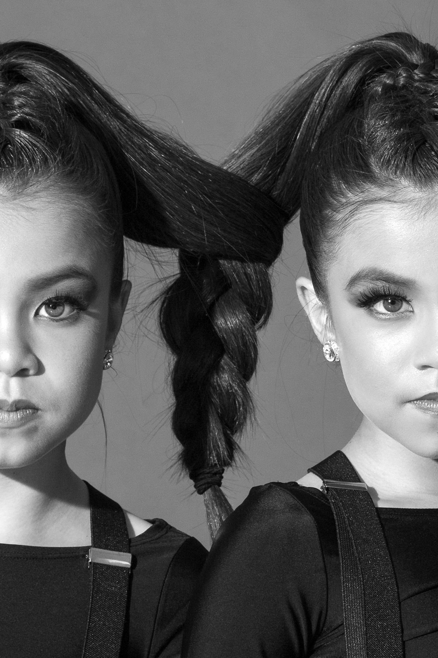 Black and white portrait of two people with their hairstyles tied together, both wearing black outfits against a plain backdrop.