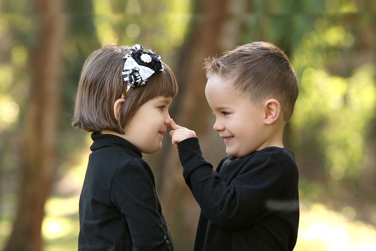 Two young children in formal black attire sharing a sweet romantic moment outdoors in a sunlit garden setting.