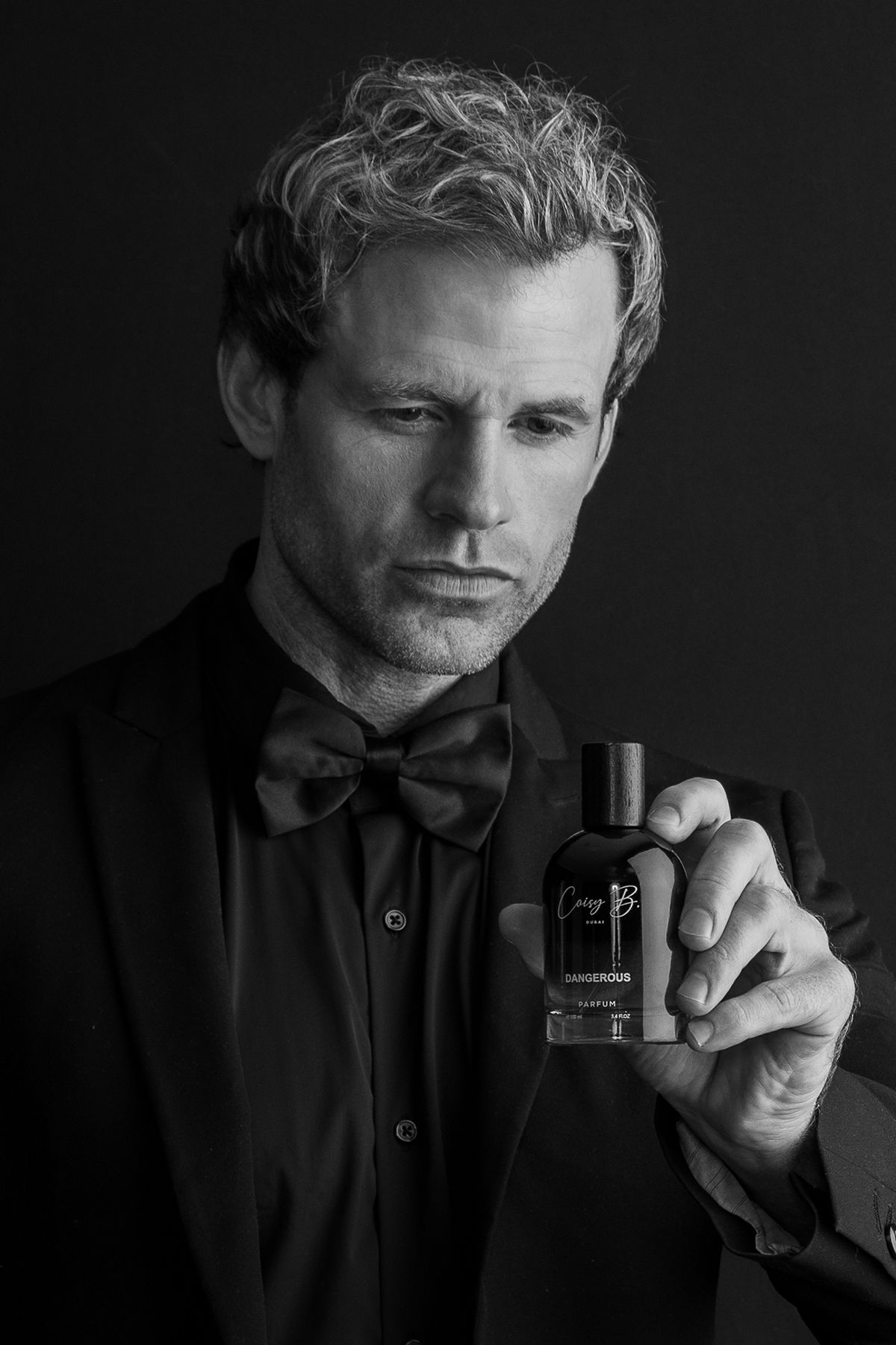 Black and white portrait of man in tuxedo and bowtie holding cologne bottle.
