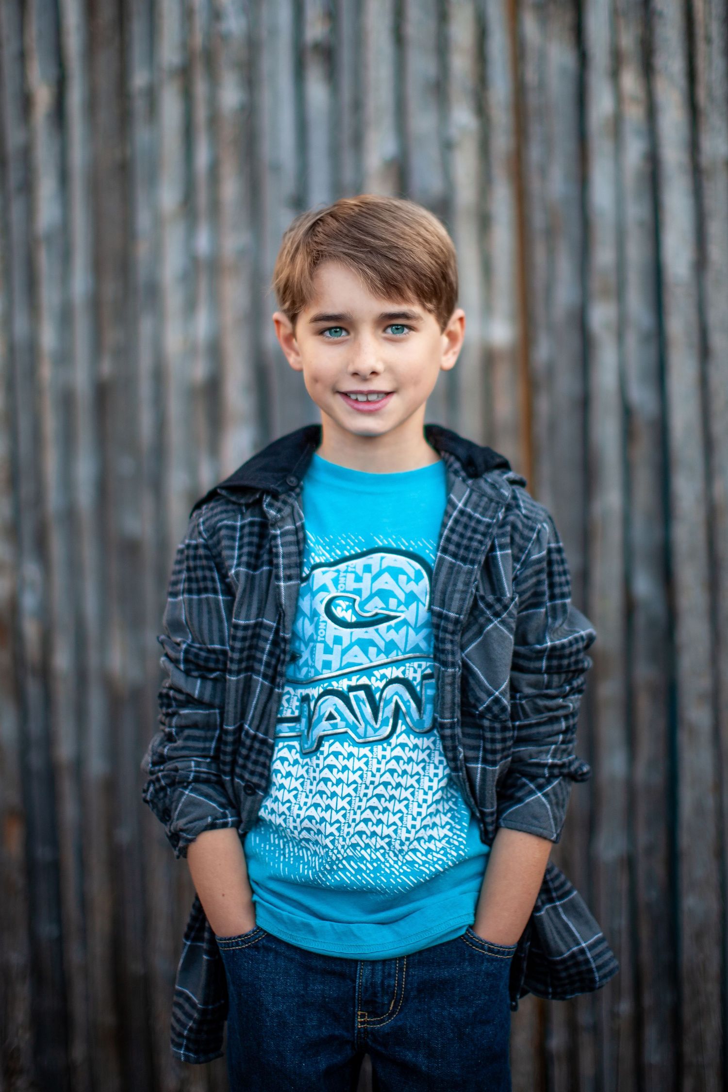 Young child wearing a bright blue graphic t-shirt and plaid flannel shirt standing against wooden slat background.