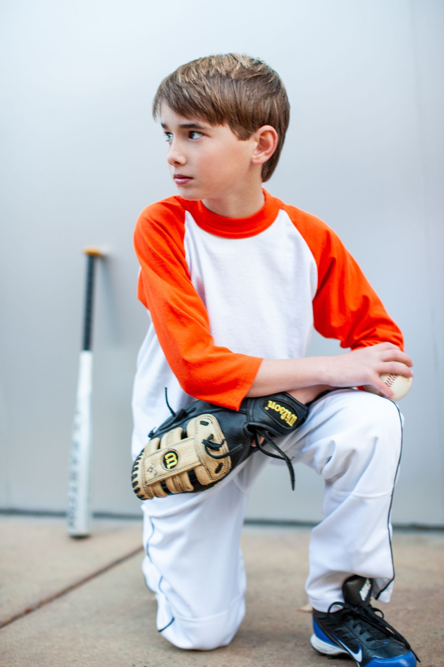 Young baseball or softball player wearing orange and white jersey kneels with catcher's mitt ready to play.
