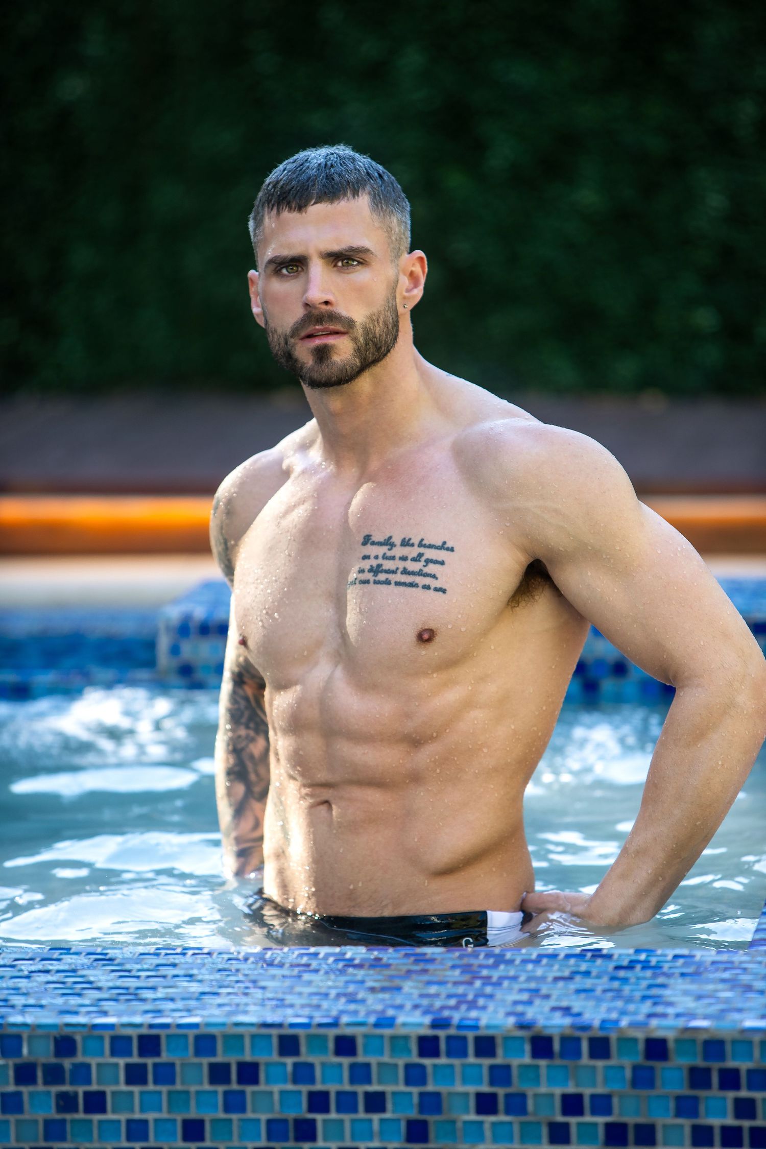 Athletic man with tattoos poses shirtless in a swimming pool with blue mosaic tile edge.