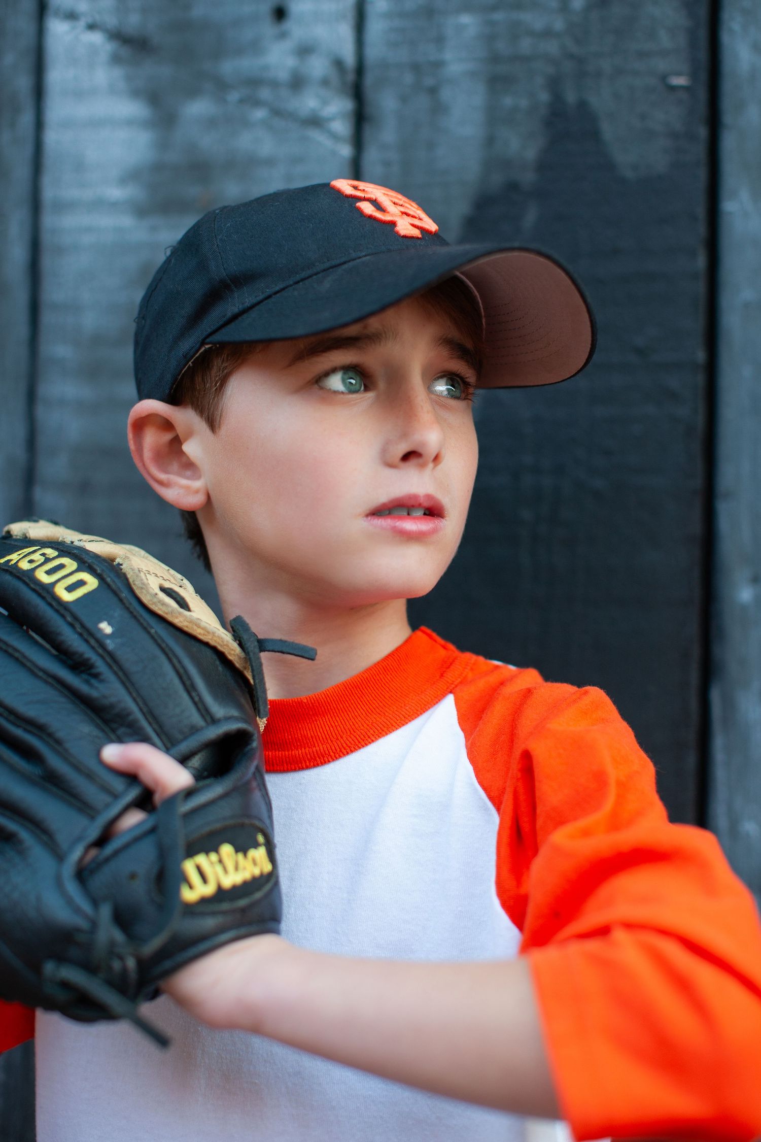 Young baseball player in orange jersey and black cap gazes upward while wearing baseball glove during practice.