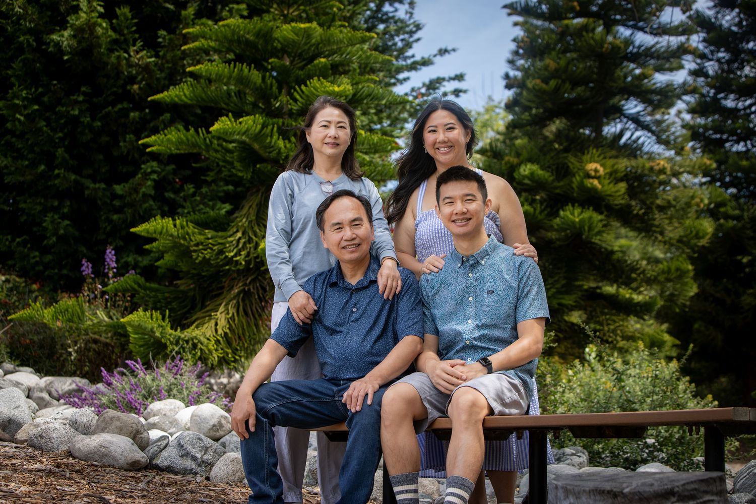 Family of four sitting on wooden bench in front of evergreen trees and garden on sunny day.