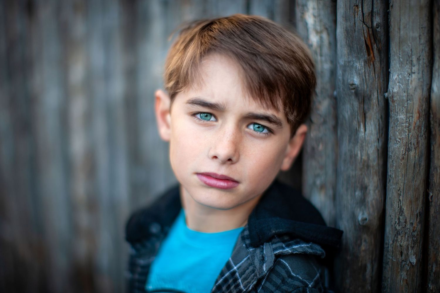 A child in a bright blue shirt poses outside against a blurred gray wooden fence background.