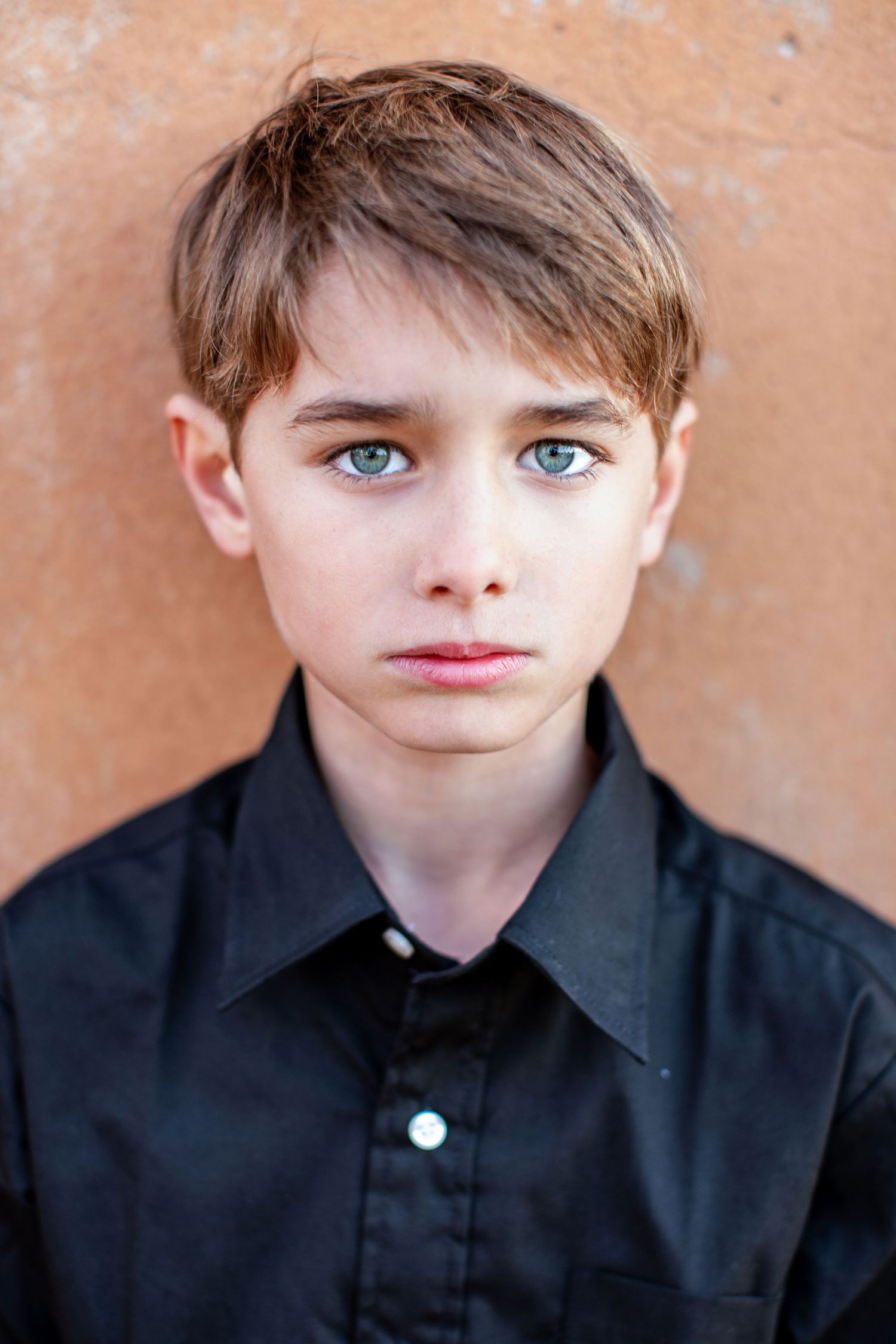 Young person in black collared shirt with striking blue eyes poses against a textured orange wall.