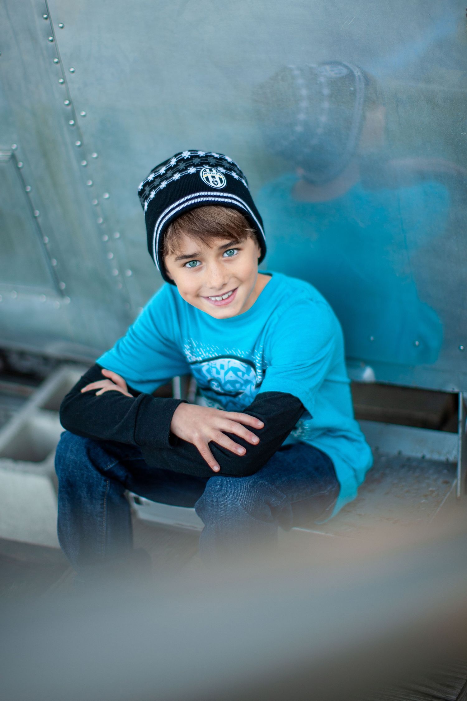 Young person in teal blue sweater and striped beanie hat sitting casually against metallic background.