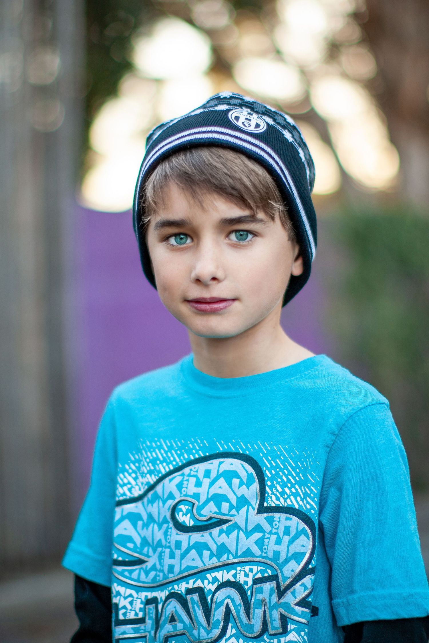 Young person in bright turquoise t-shirt and dark cap smiling in outdoor setting with blurred background.