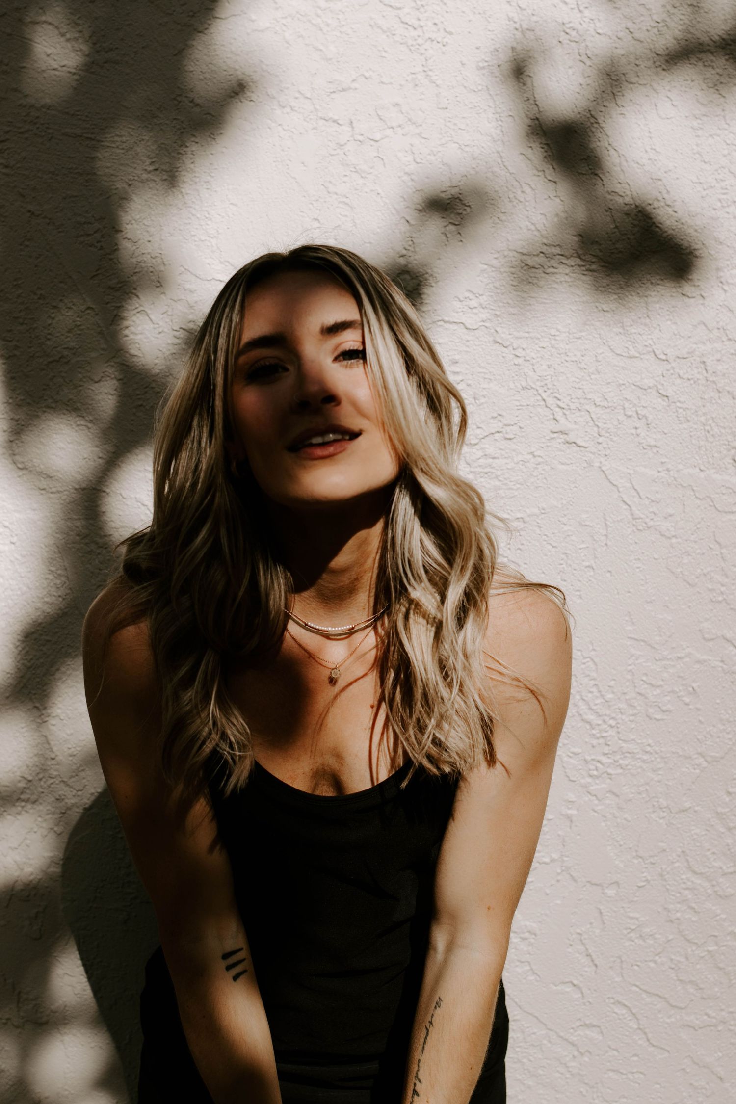 Blonde woman in black top poses against textured white wall with leaf shadows in natural sunlight.