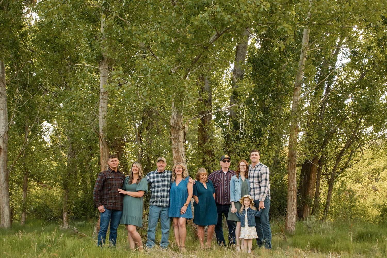Family of eight posing outdoors in a lush green meadow surrounded by tall trees during golden hour.