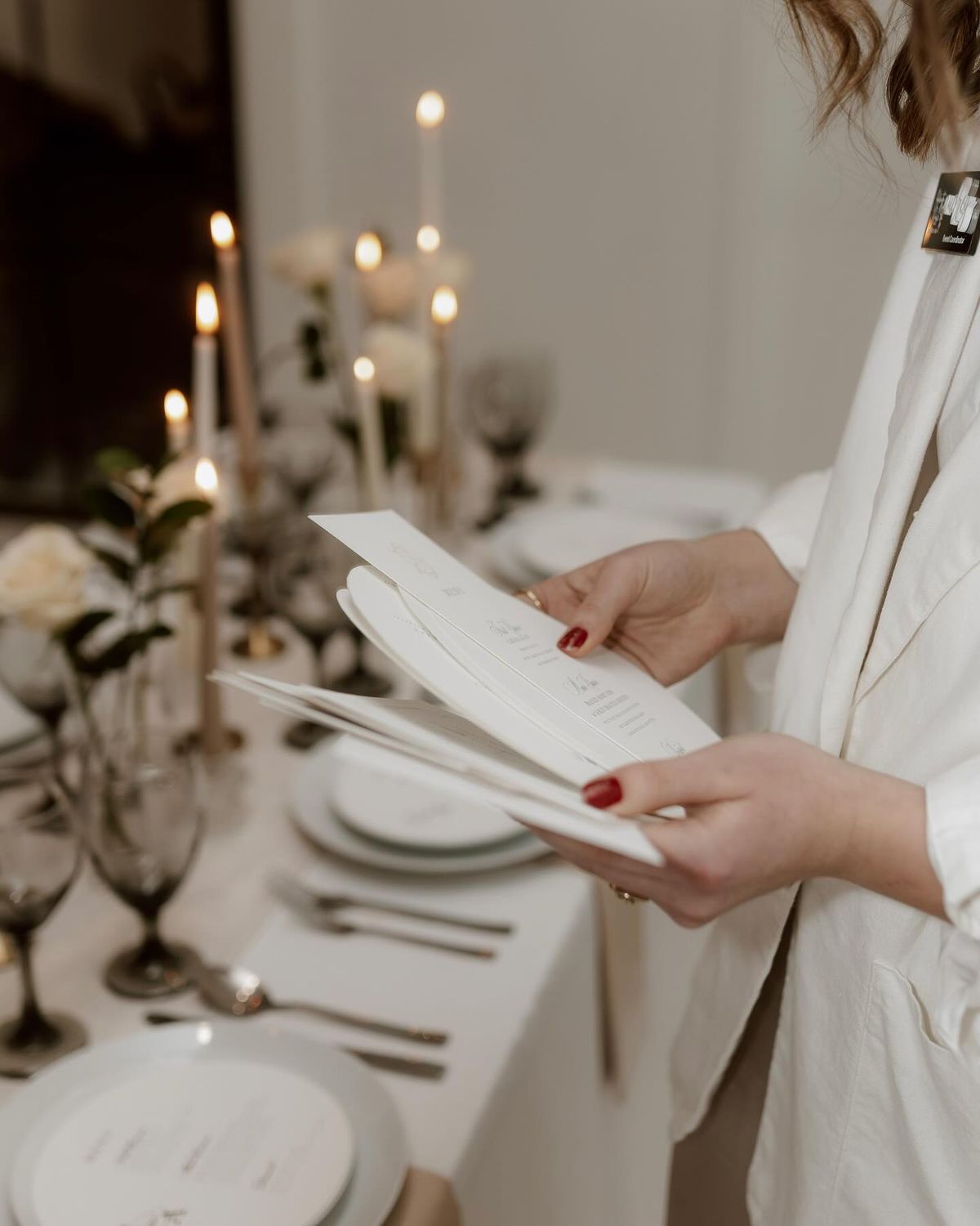 Woman in white linen holding menu cards at an elegant candlelit dinner table setting with floral decor.