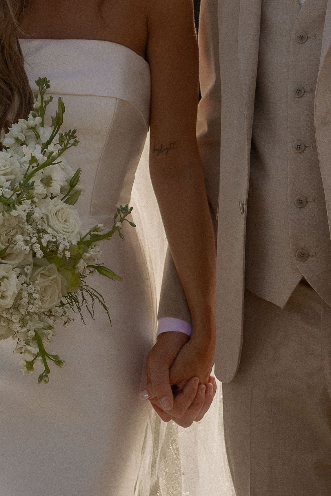 Bride and groom holding hands, bride in white strapless gown holding white floral bouquet, groom in beige suit vest.