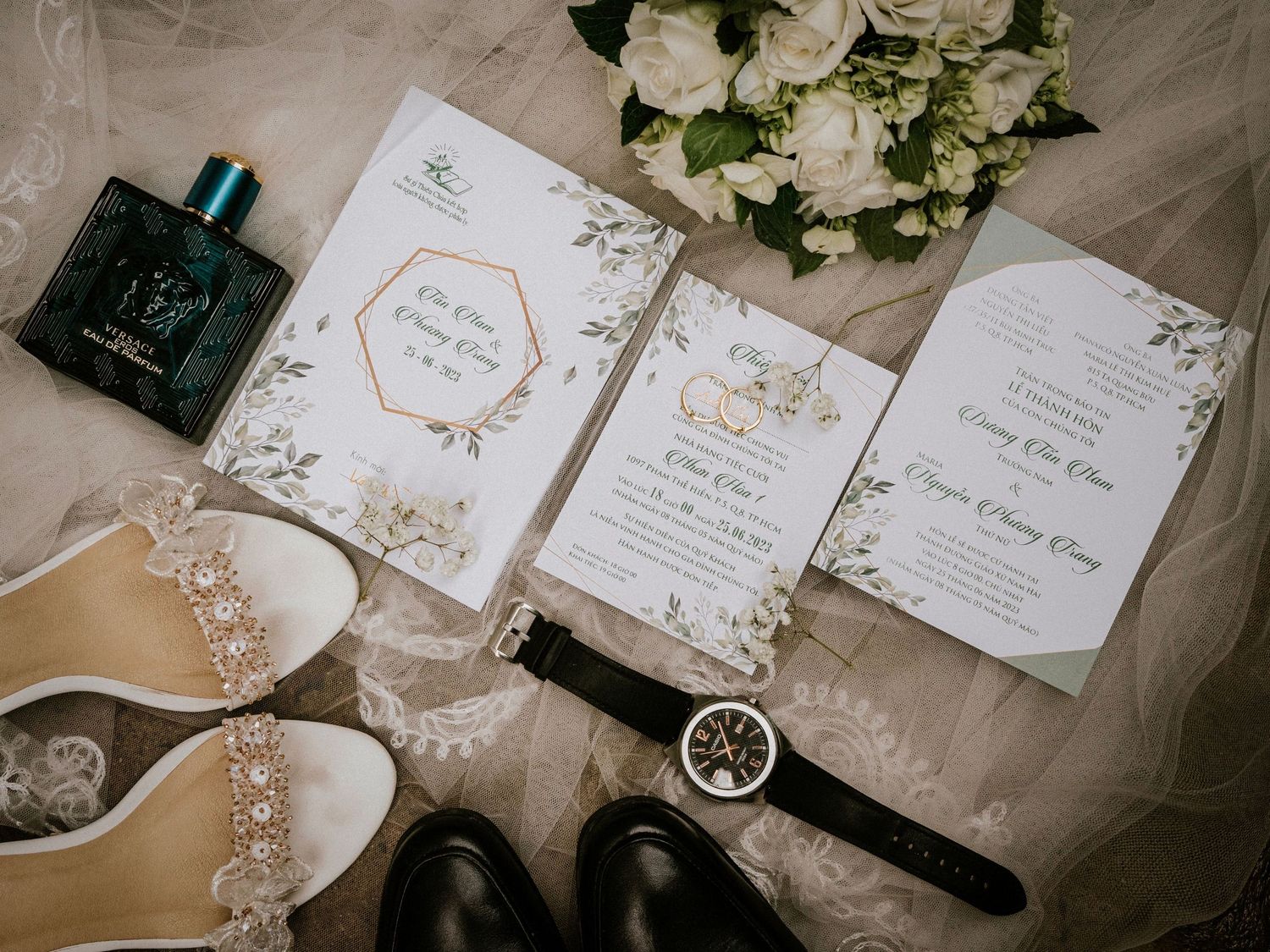 Elegant wedding flat lay featuring invitations, white roses, bridal heels, watches, and cologne on lace fabric.