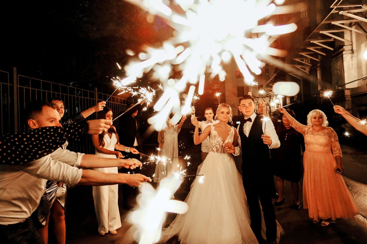 Bride and groom celebrate with sparklers surrounded by guests at a romantic nighttime wedding reception.
