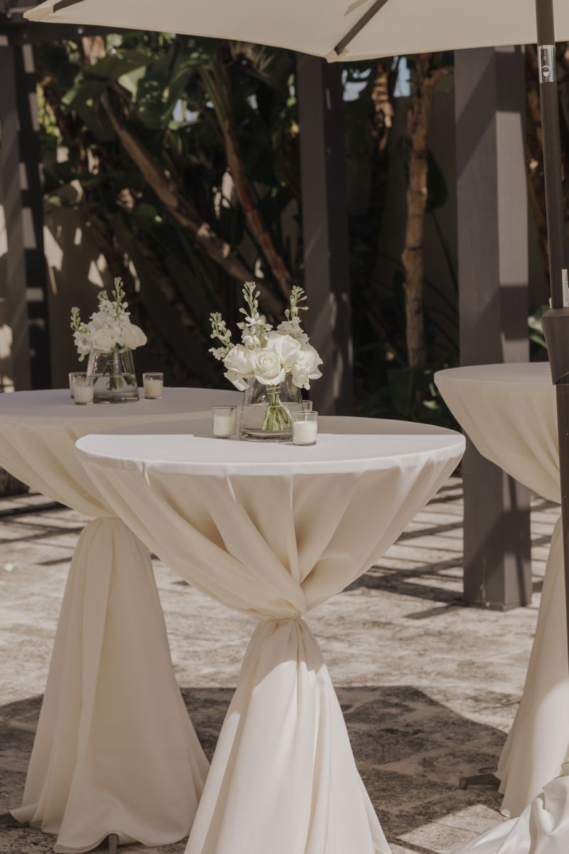 Elegant outdoor cocktail table with white draping and floral centerpiece at a wedding reception.