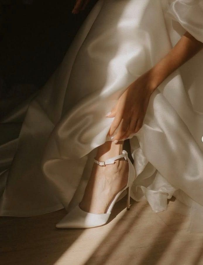 Bride adjusting white satin ankle-strap heels beneath flowing wedding gown in warm natural light.