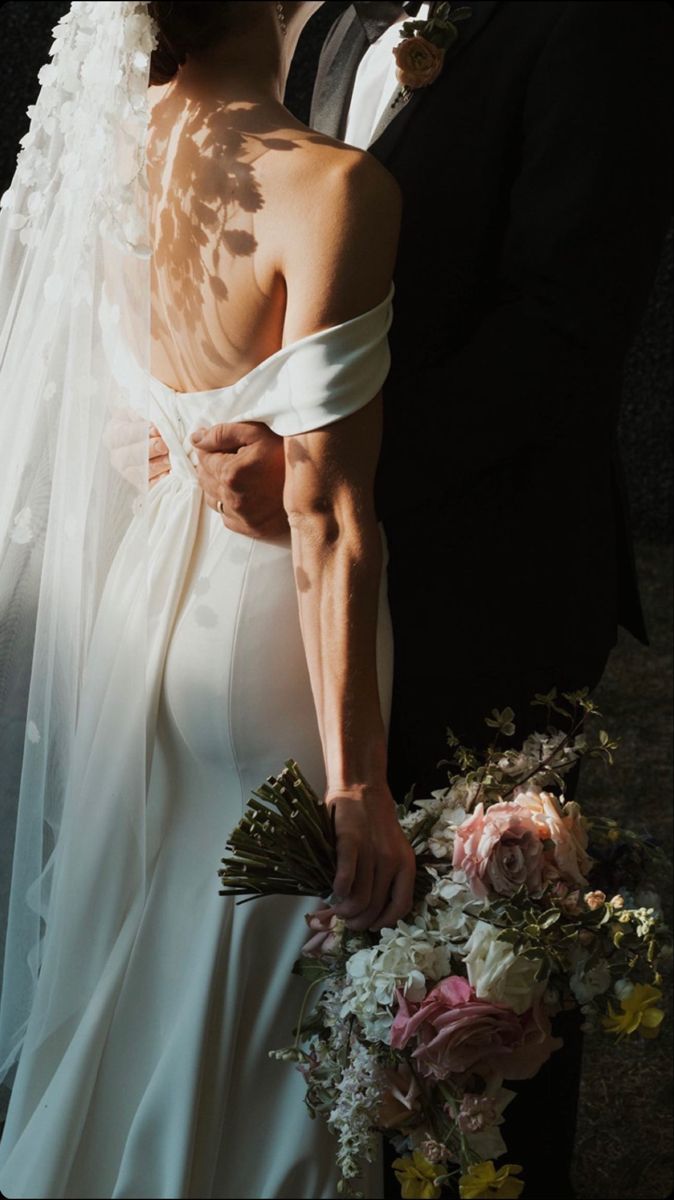 Bride in backless white gown holding floral bouquet stands with groom in black suit at wedding ceremony.