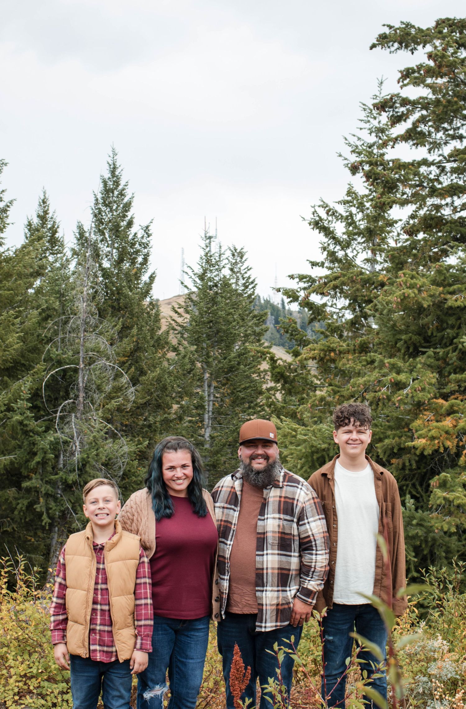 Family of four smiling outdoors among evergreen trees and autumn foliage in a mountain forest setting.