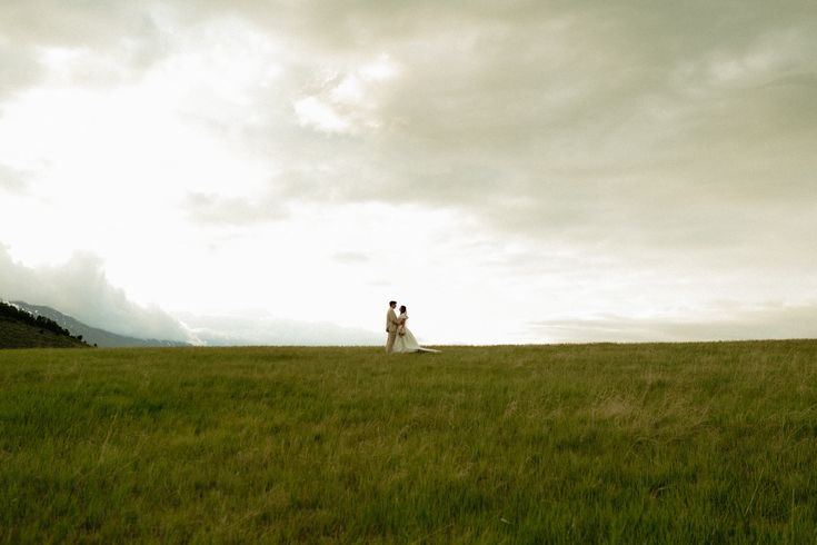A couple stands in a vast green meadow under dramatic cloudy skies, capturing a romantic wedding moment in nature.