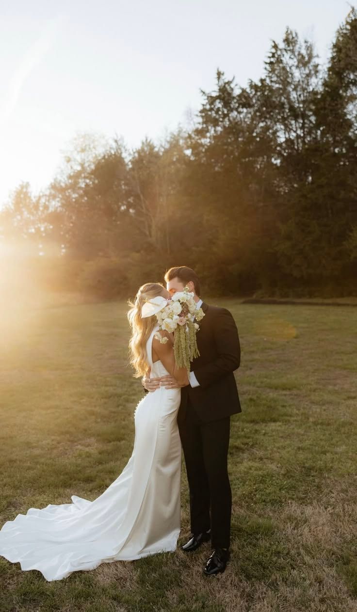 A bride and groom share a kiss at golden hour on a lush green lawn, the bride holding a white floral bouquet.