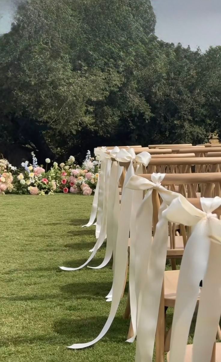 Outdoor wedding ceremony aisle with wooden chairs adorned with white satin ribbon bows, leading to a floral arch backdrop.