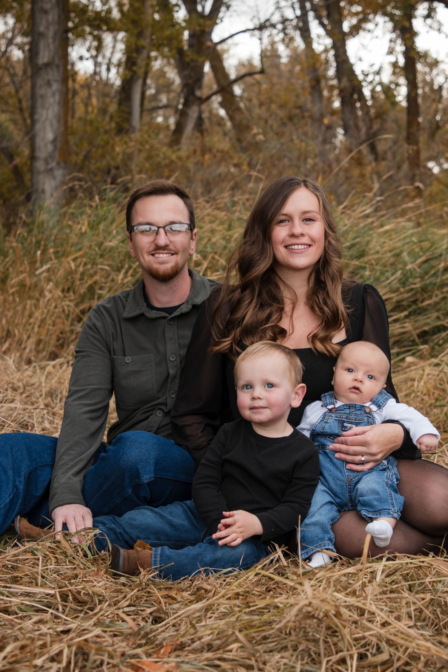 A smiling family of four poses outdoors on hay, with two parents and two young children amid autumn trees.