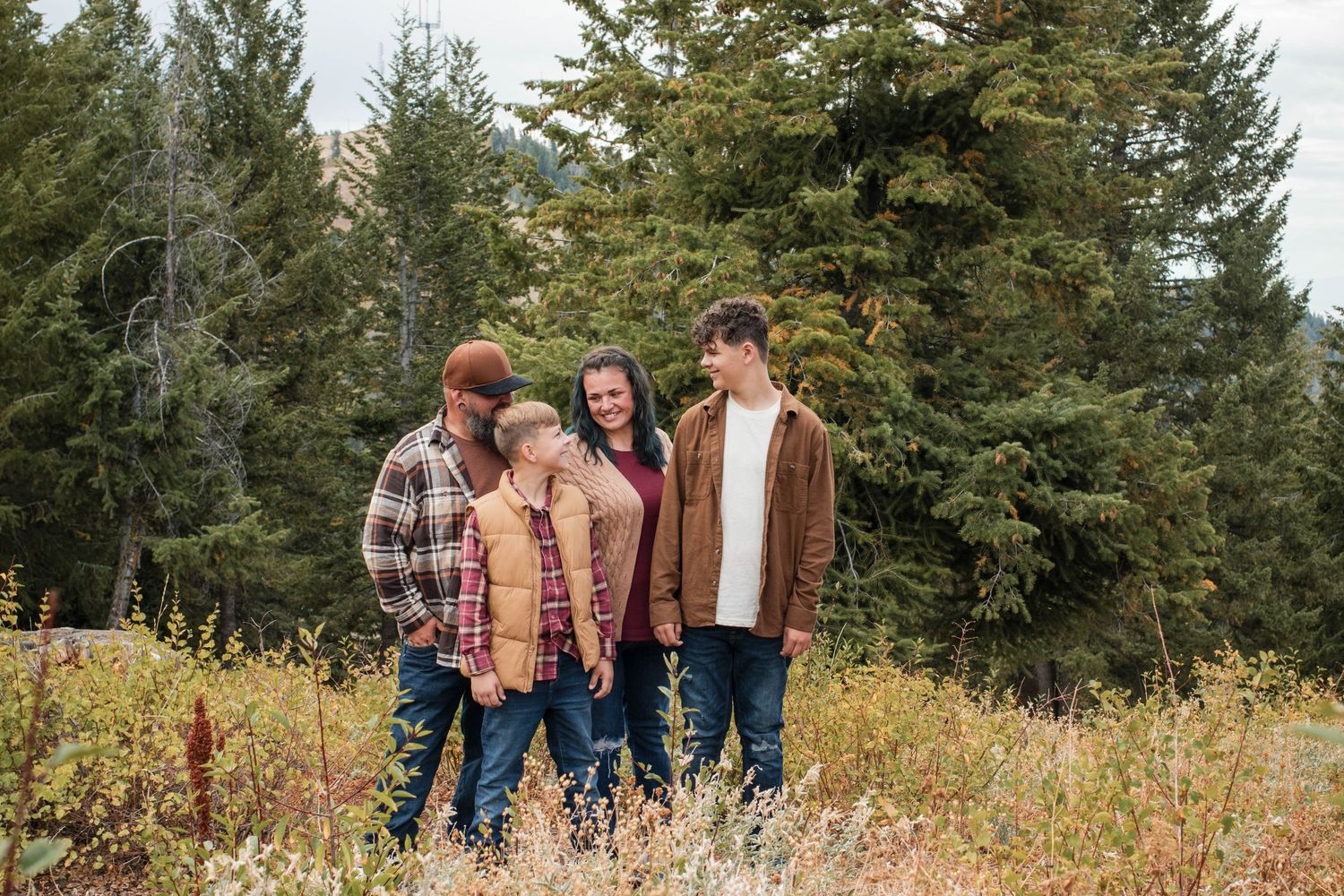 Family of four posing outdoors in autumn field with evergreen trees, wearing coordinated fall outfits.