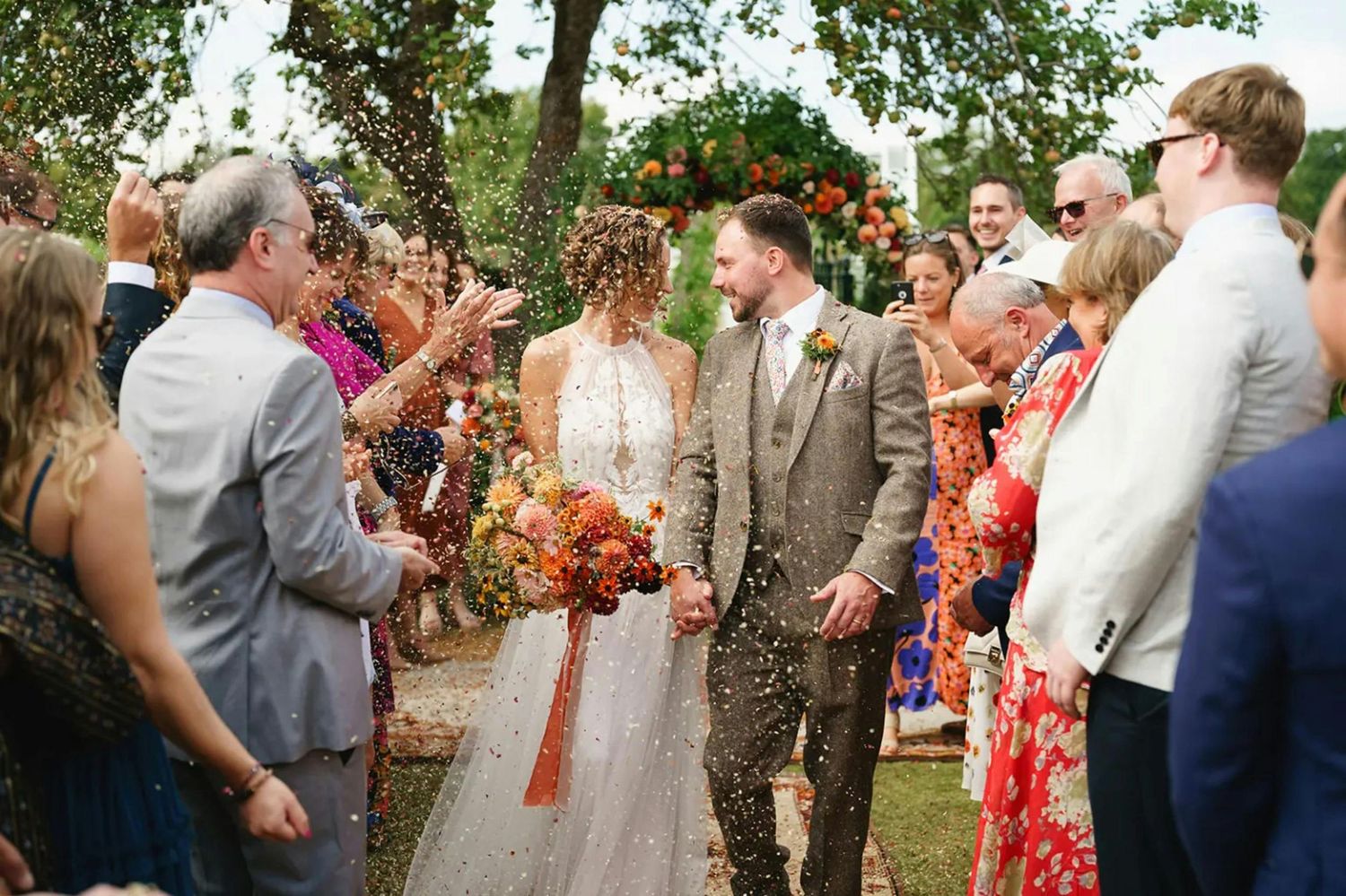 Bride and groom walk through confetti shower surrounded by smiling wedding guests outdoors.