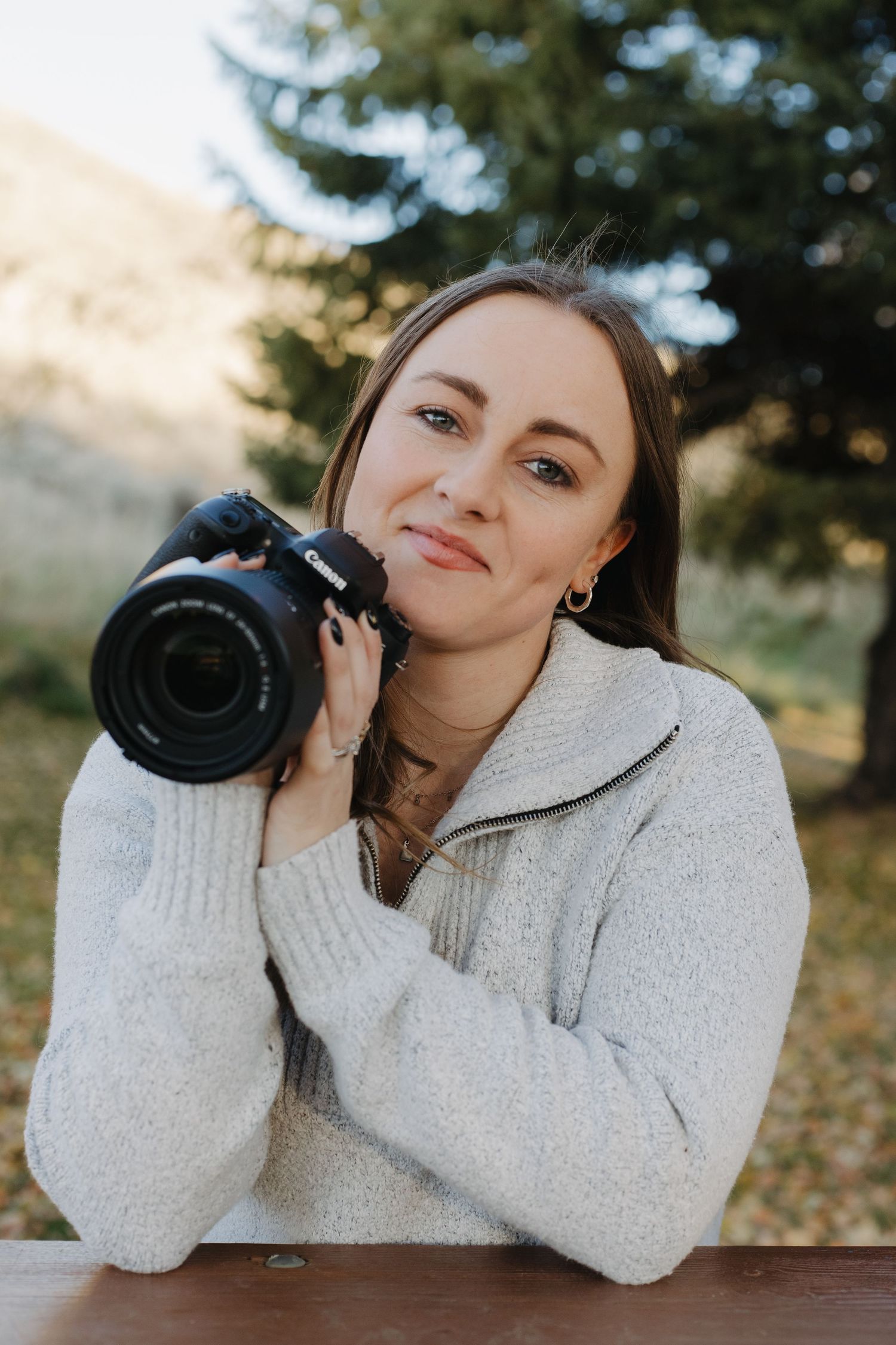 Woman in white knit sweater holding a Canon camera outdoors with trees in background.