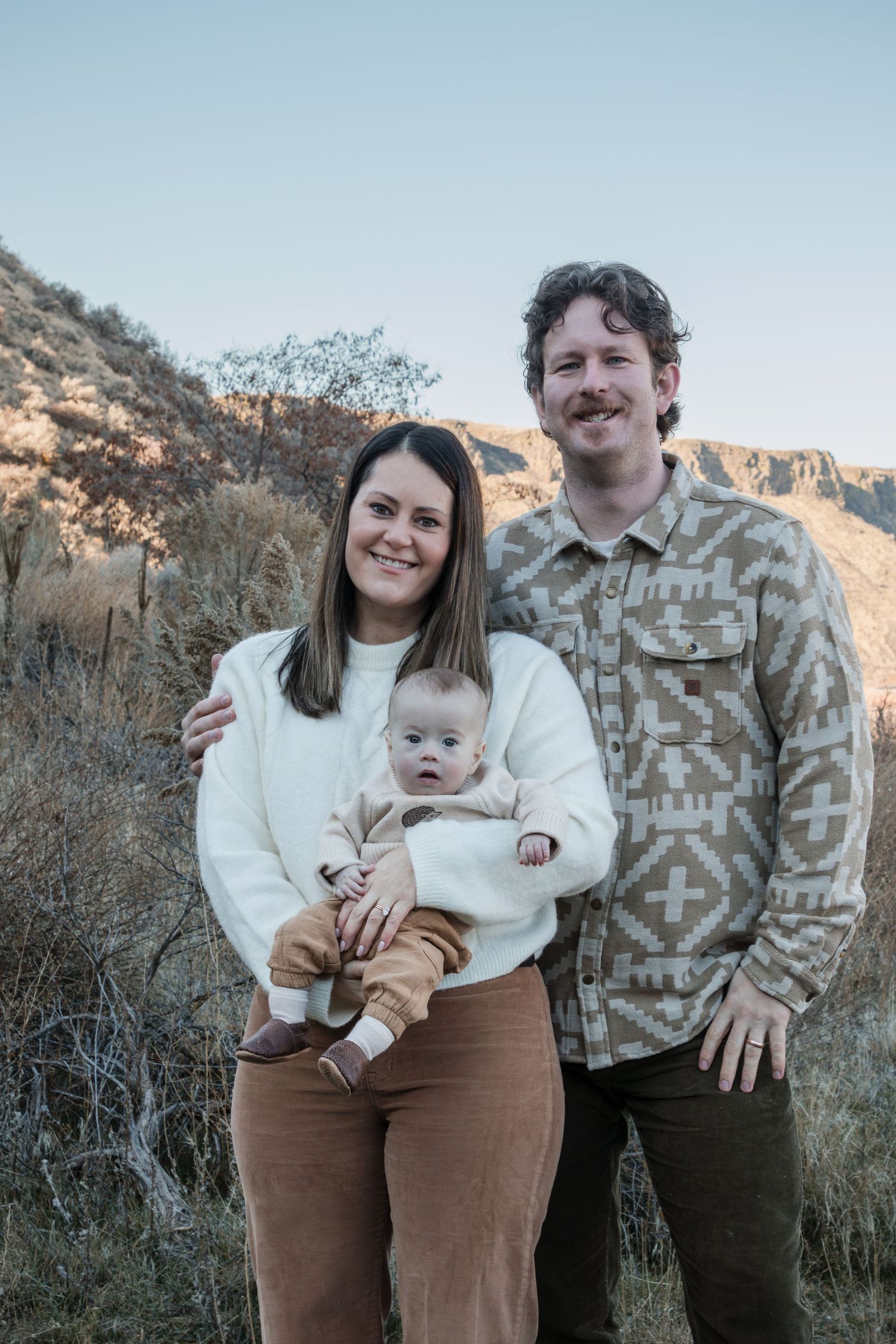 A smiling couple poses outdoors holding their baby, with desert mountains in the background.