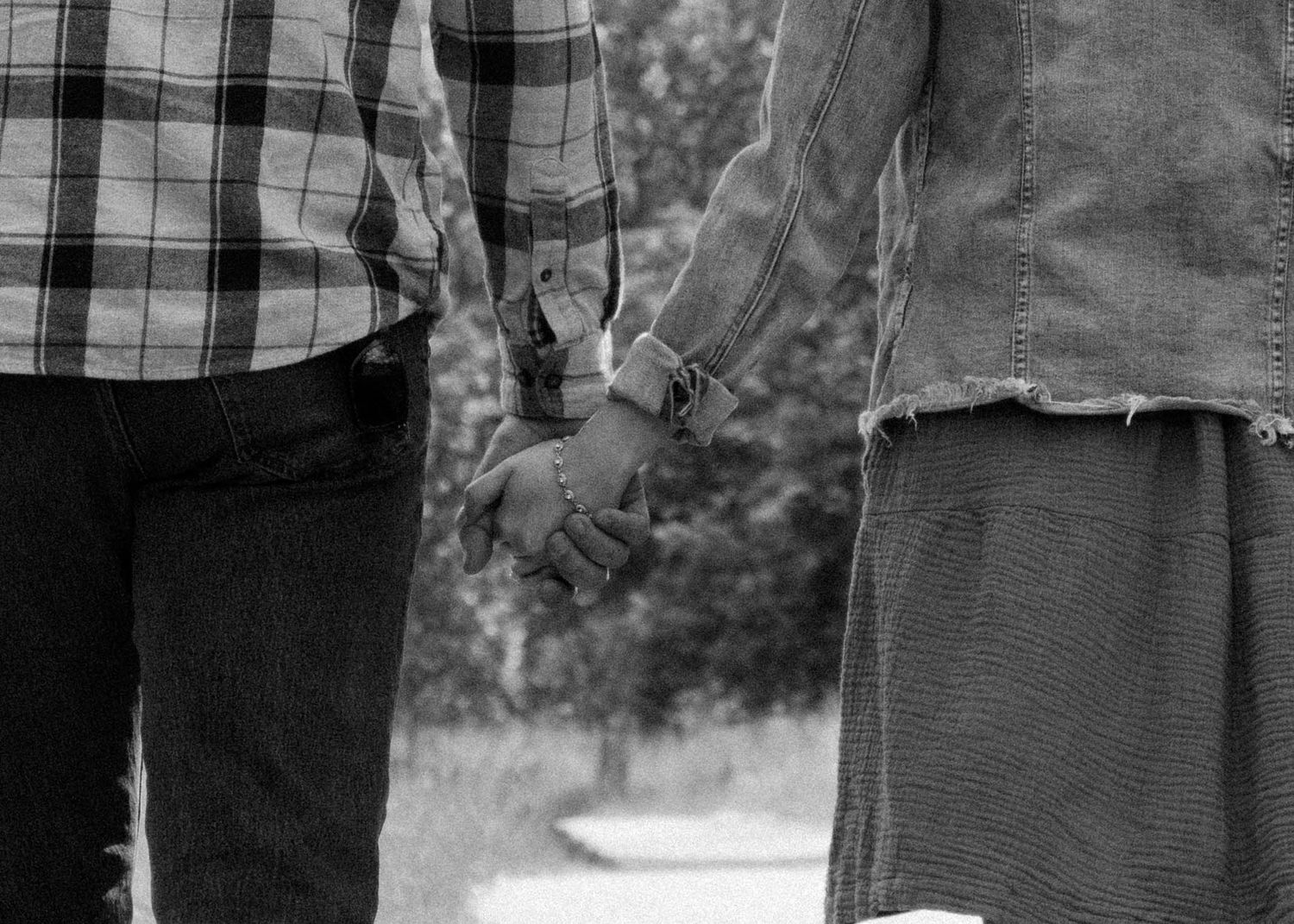 Black and white photo of a couple holding hands, wearing casual clothes, with blurred trees in the background.