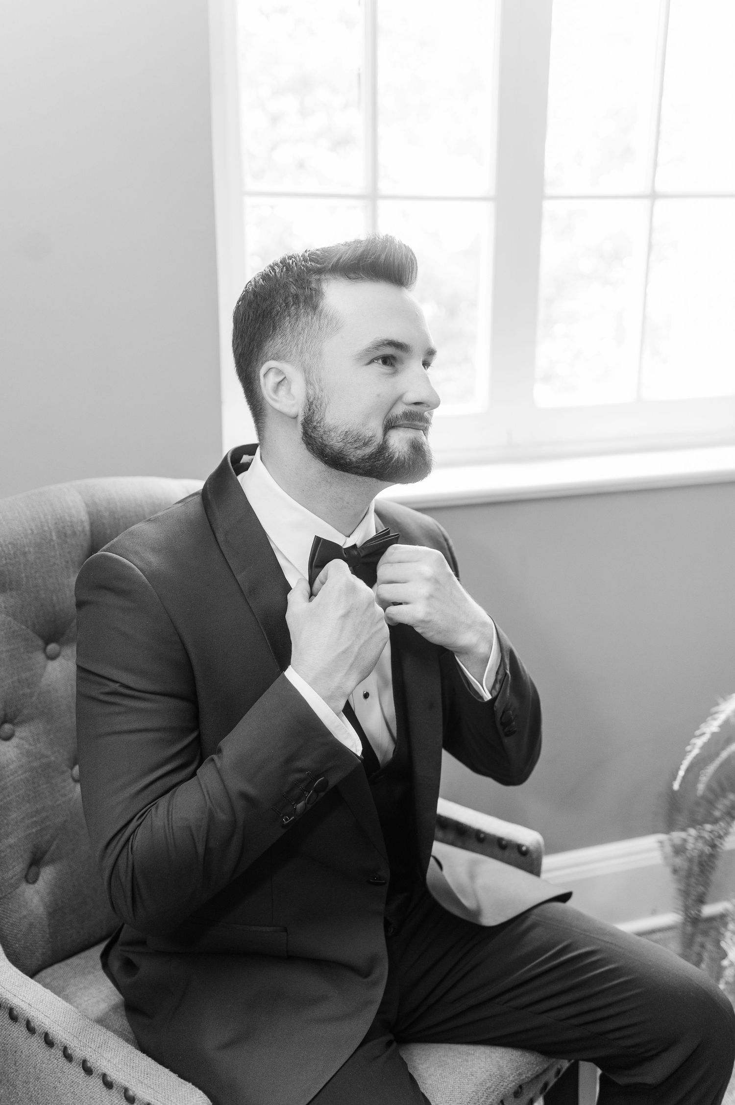 Groom in black suit sits thoughtfully in vintage chair by window.