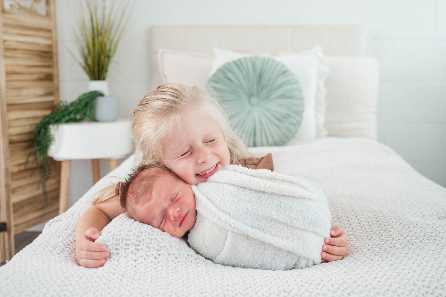 Smiling toddler hugging swaddled newborn baby on white bed in bright nursery room.