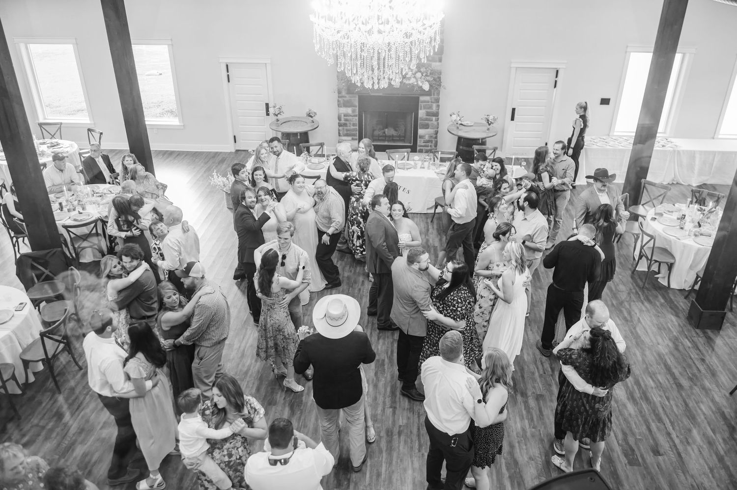 Black and white overhead photo of wedding guests mingling on a wood dance floor beneath a chandelier in a rustic reception hall.