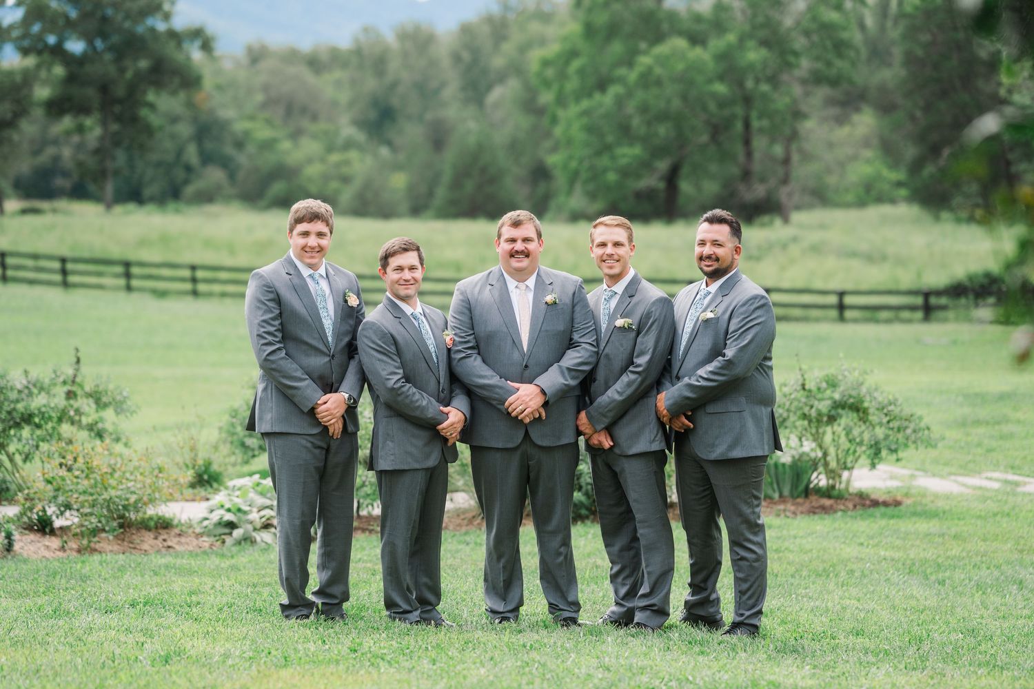 Five groomsmen in matching gray suits smile together on a green lawn at an outdoor wedding venue.