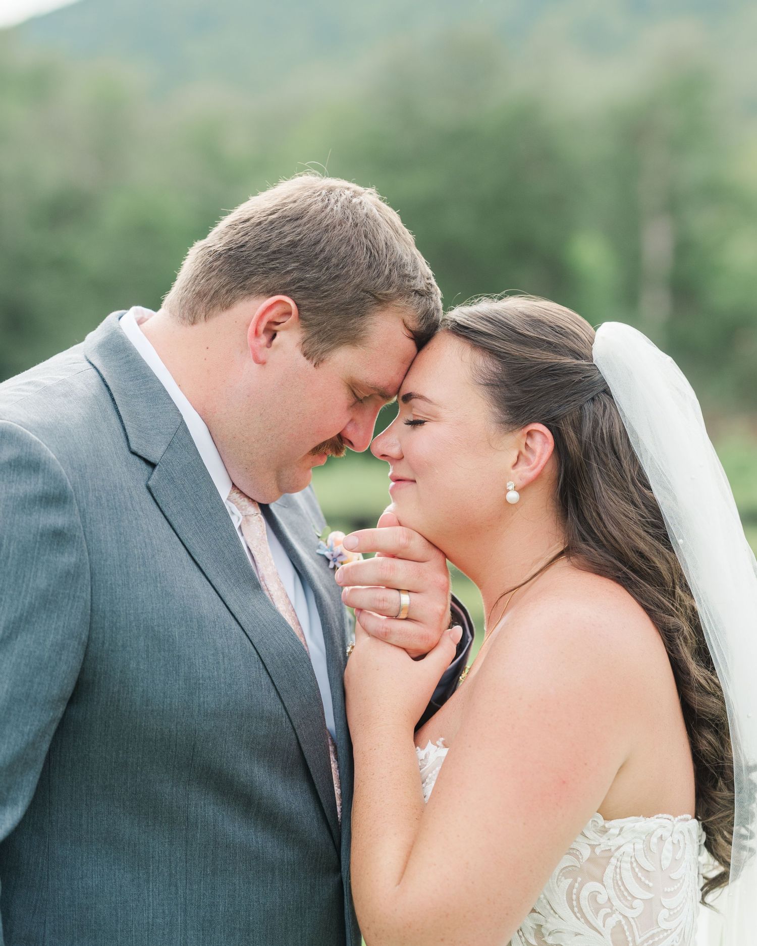 A bride and groom share a romantic forehead touch, holding hands in wedding attire with a scenic mountain backdrop.
