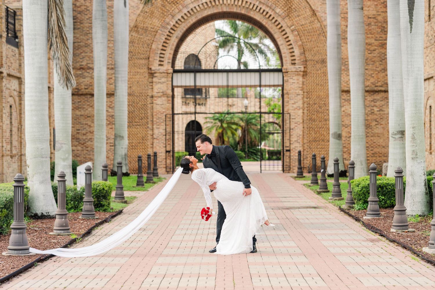 Bride and groom share romantic dip kiss on brick walkway framed by ornate arched building and palm trees in Texas