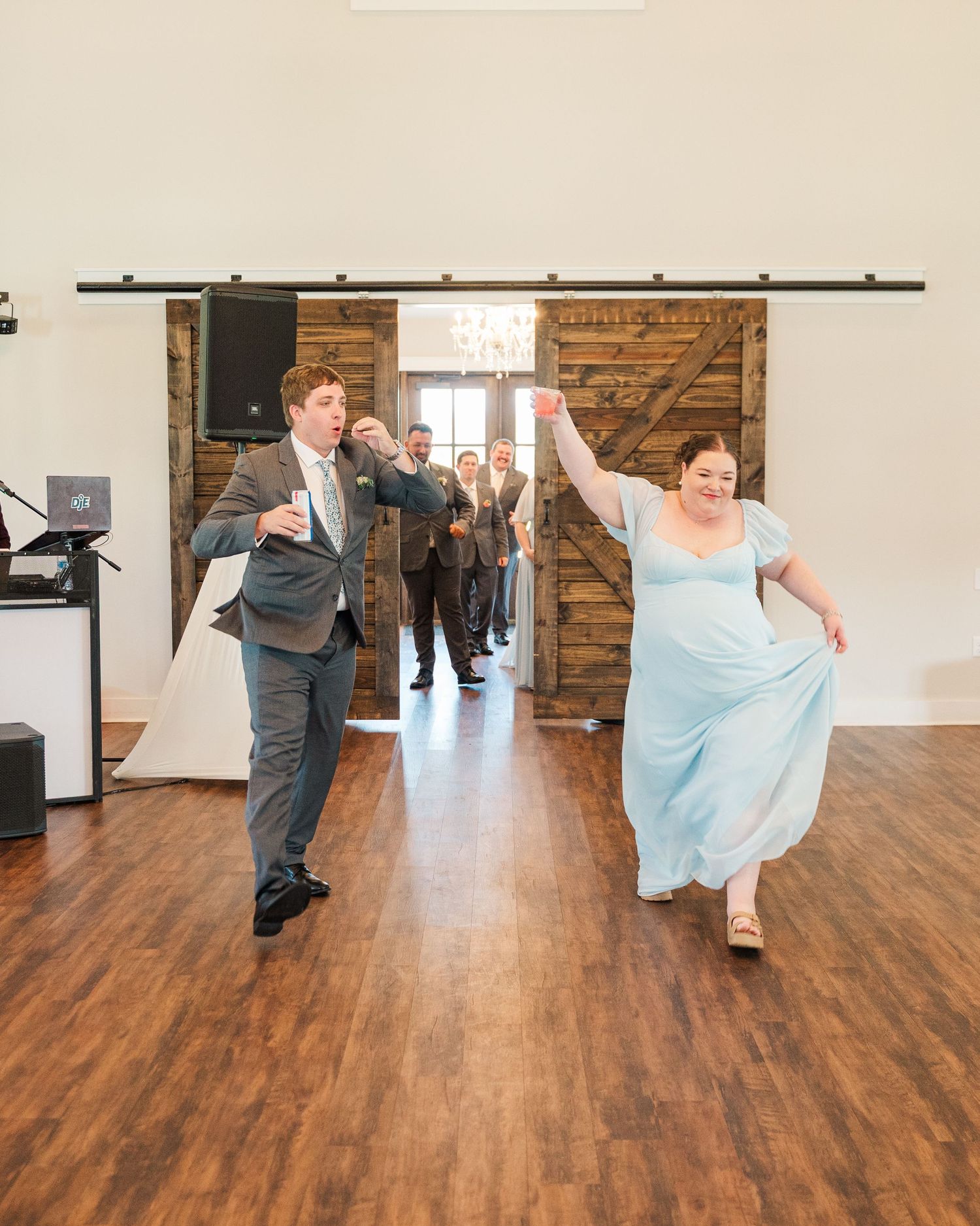 Bridesmaid in light blue gown and groomsman make a lively entrance through rustic barn doors at reception.