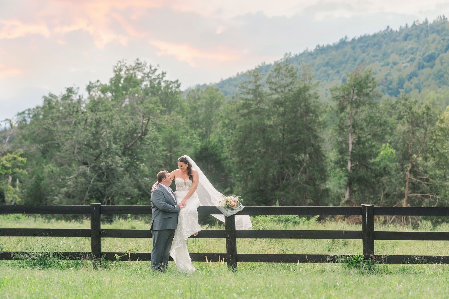 Bride and groom share a romantic moment in a green meadow with mountain views and wooden fence at sunset.