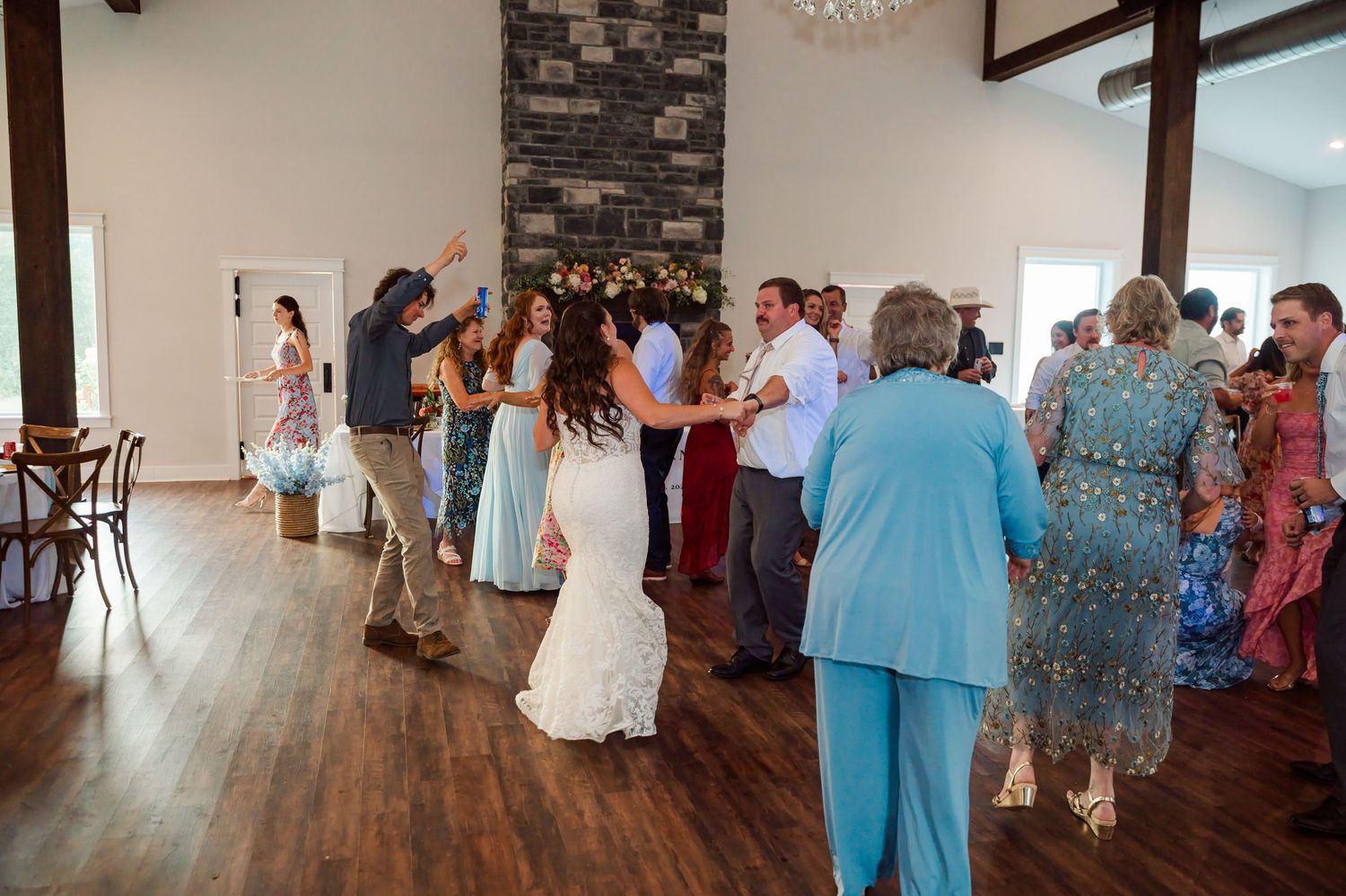 Bride in a white wedding gown dances with guests on a wood floor in a rustic venue with a stone fireplace and large windows.