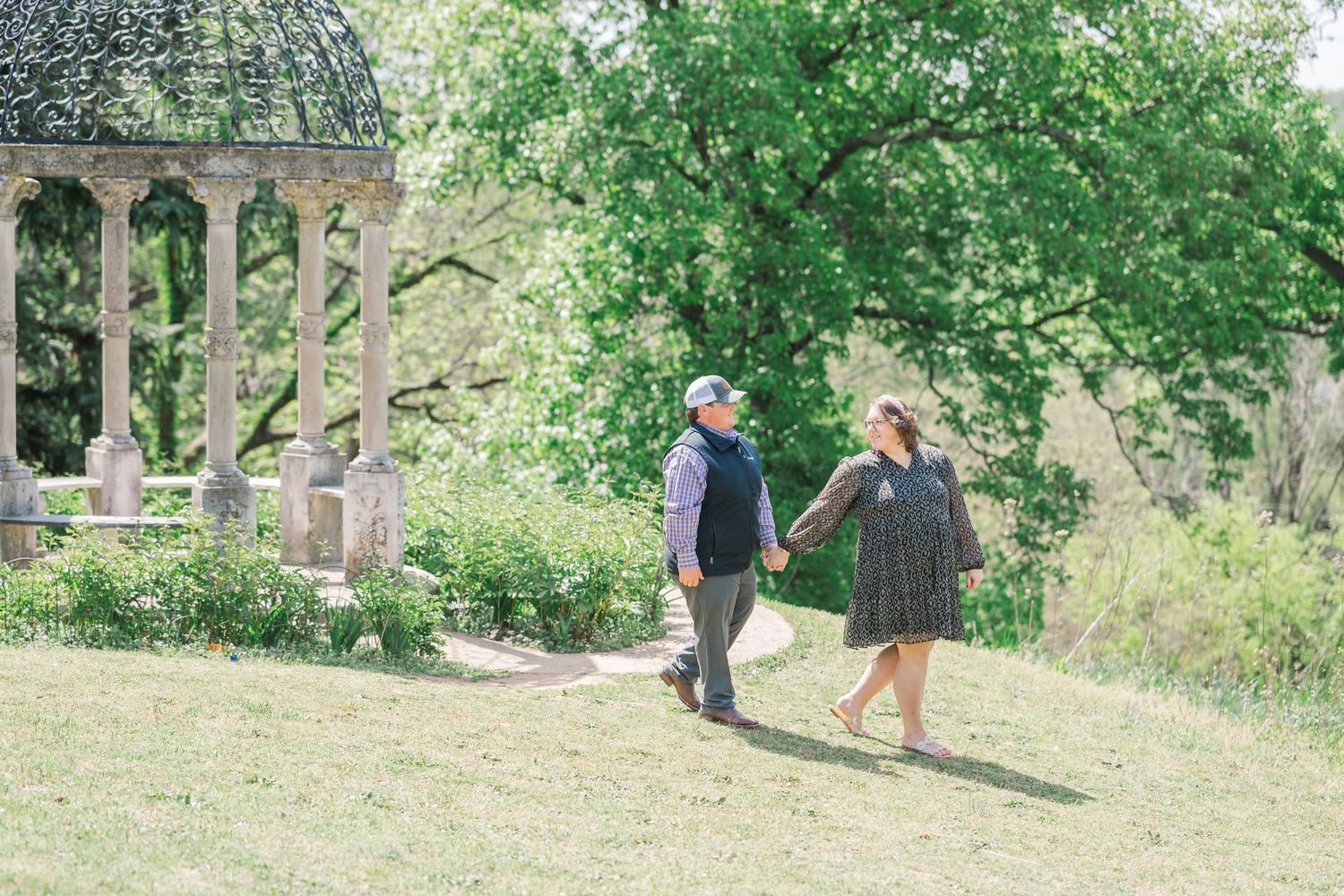 Couple holding hands walking through a lush green park with a stone gazebo and blooming trees in the background.
