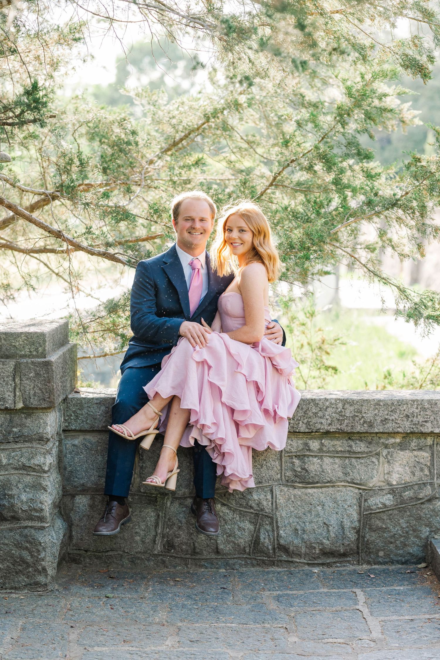 Couple in pink dress and navy suit sitting on stone wall outdoors, smiling, surrounded by lush green trees.