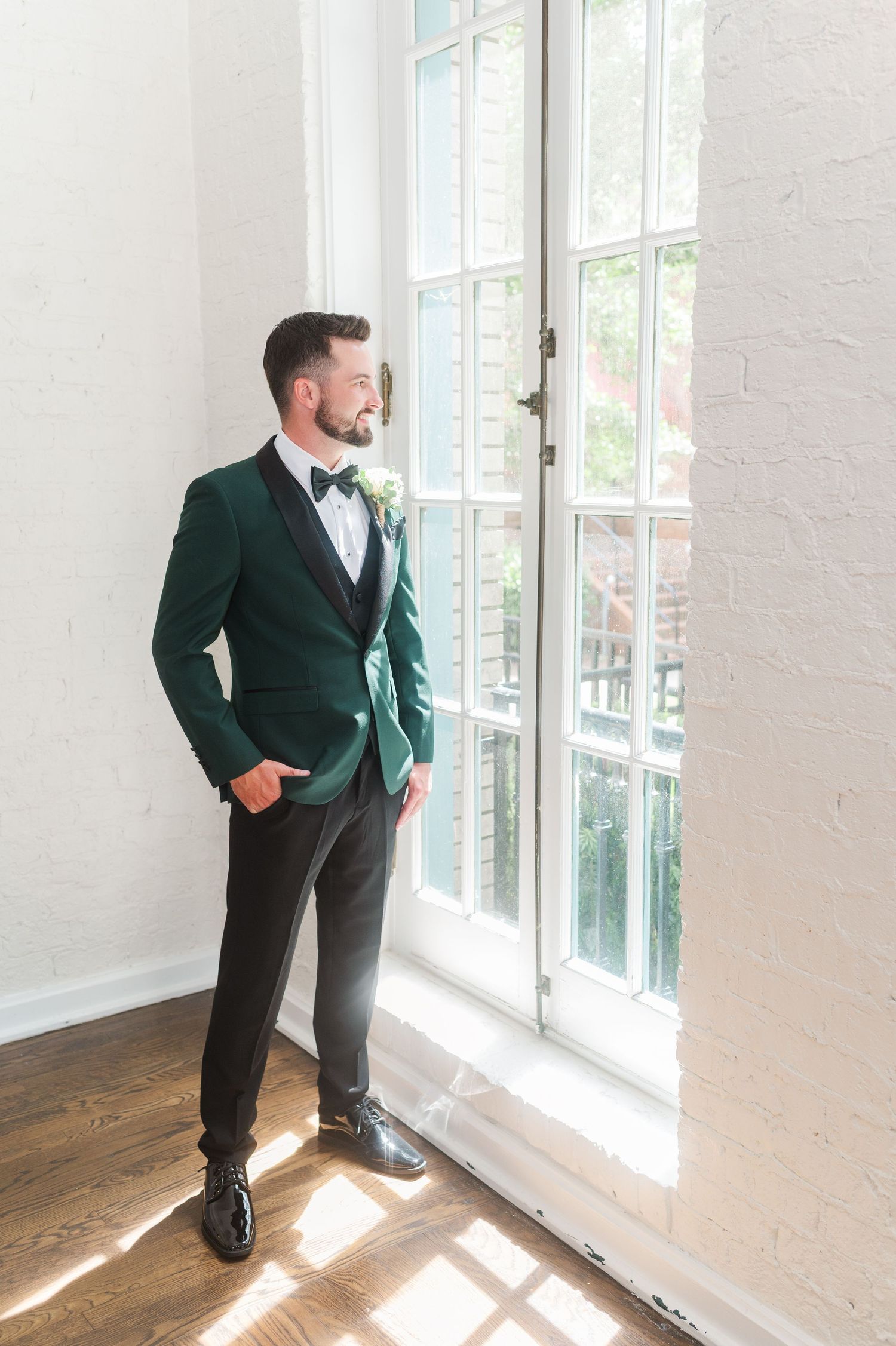 Groom in emerald green jacket poses by bright windows in formal setting.