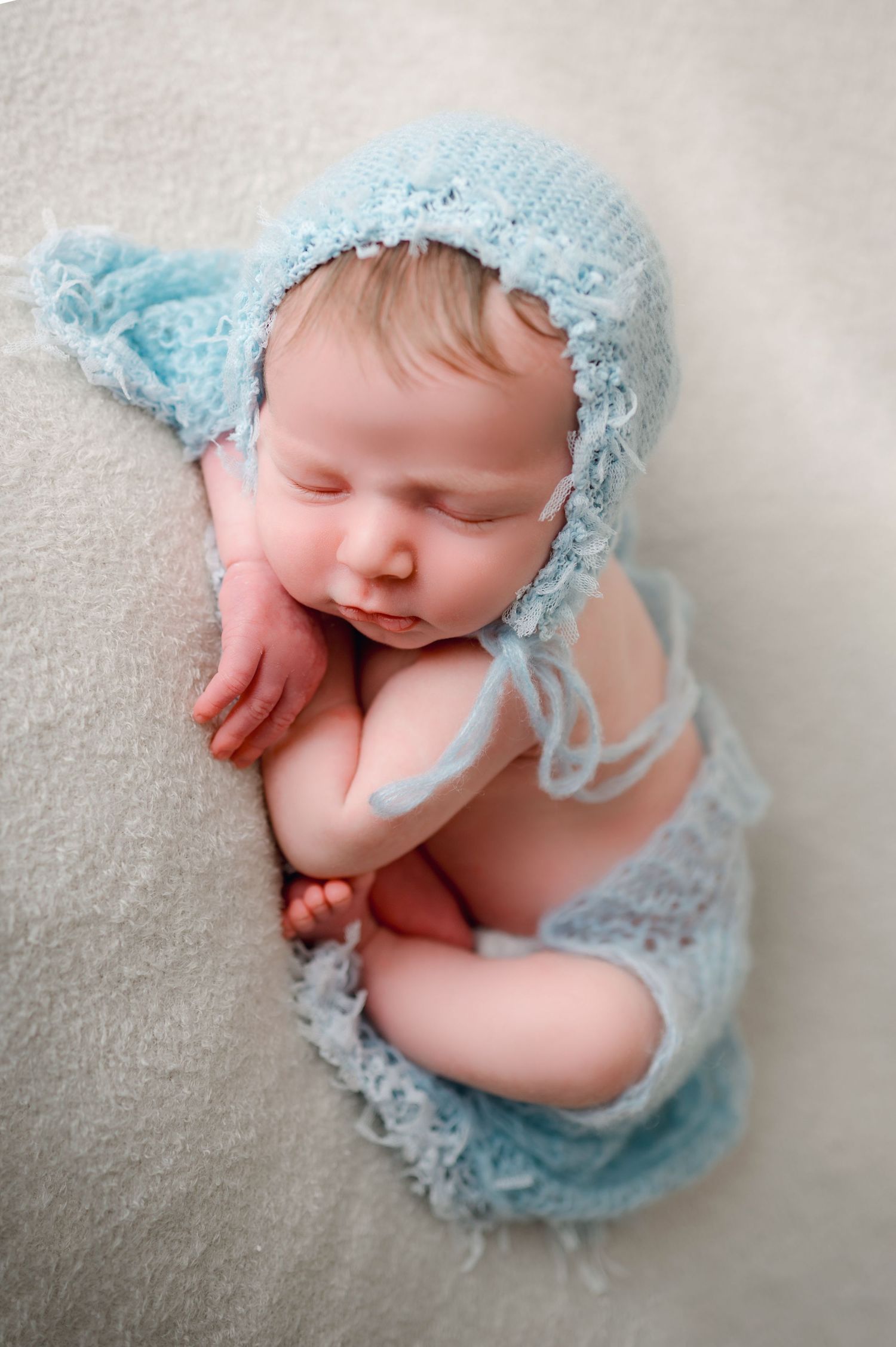 Sleeping newborn wrapped in soft blue lace bonnet on white blanket.