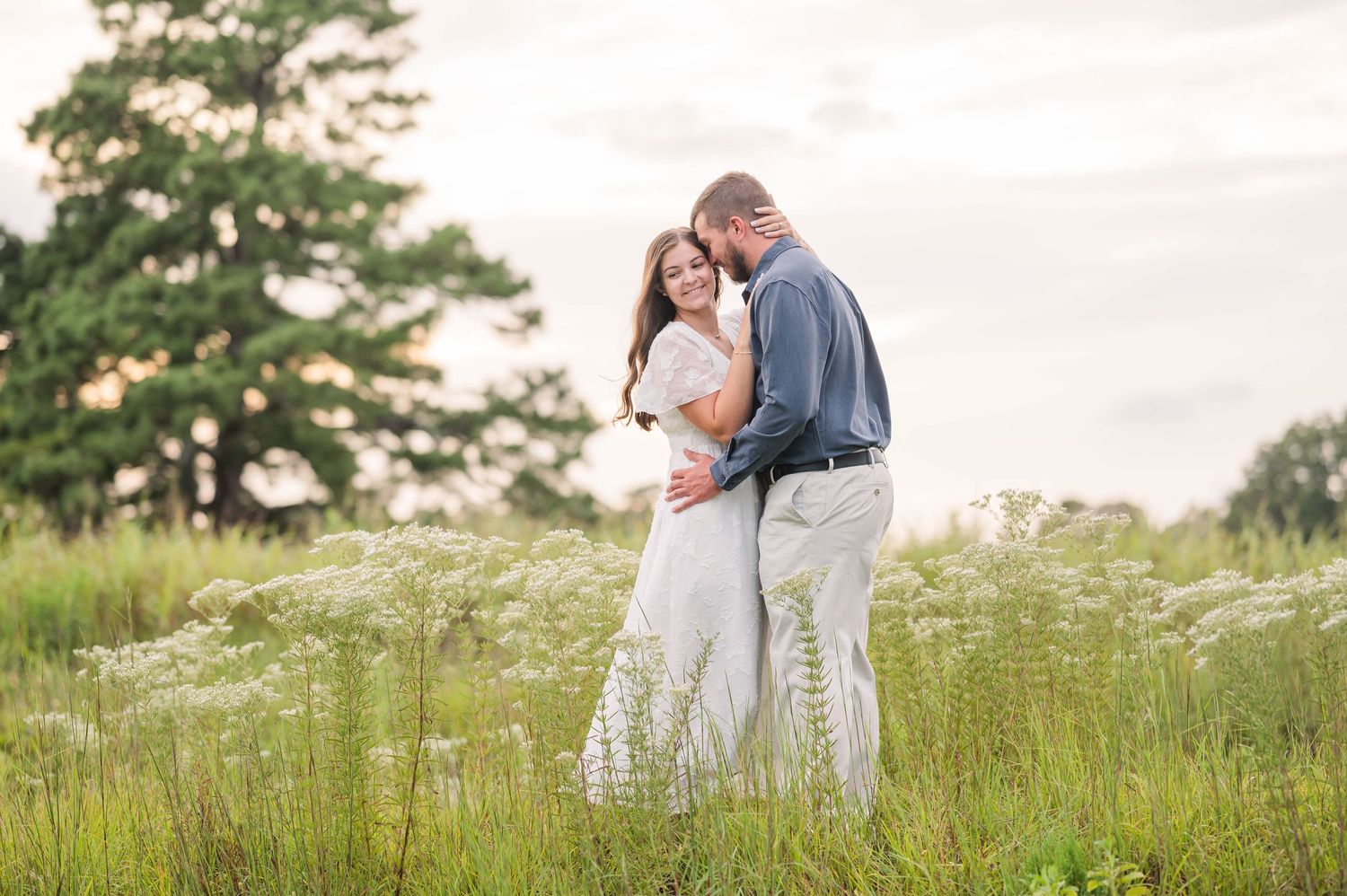 Couple embracing in a wildflower meadow with a large tree backdrop during golden hour engagement photo session.